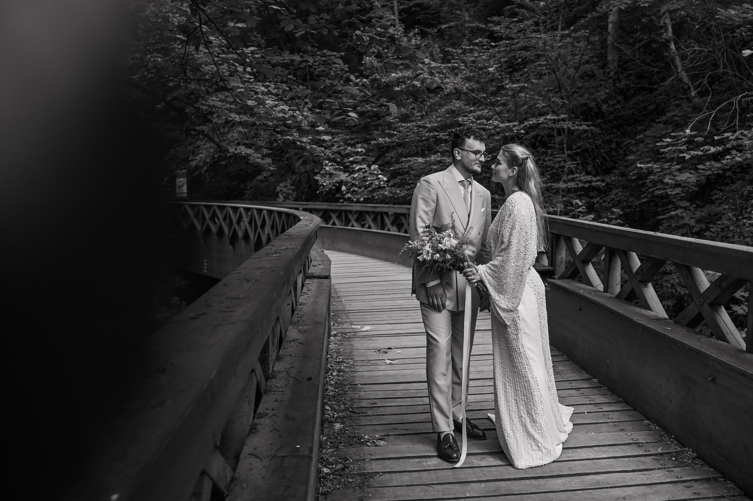 A black-and-white photo of a bride and groom standing close on a wooden bridge surrounded by trees, holding hands, and gazing into each other's eyes. The groom is wearing a light-colored suit and glasses, and the bride is wearing a long dress with a 