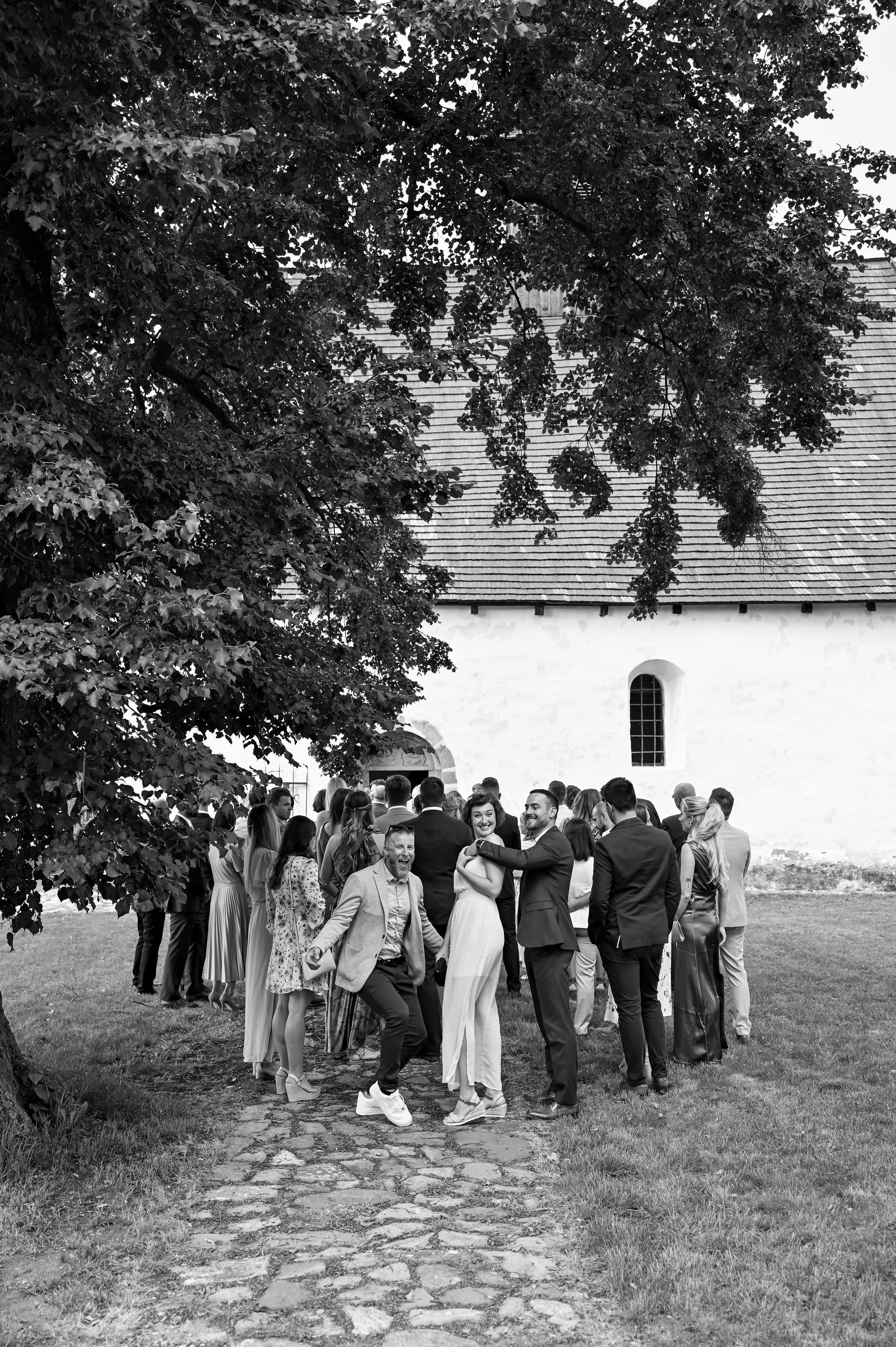 A group of people dressed in formal attire gather outdoors on a cobblestone path near a white building, smiling and socializing under a large tree.