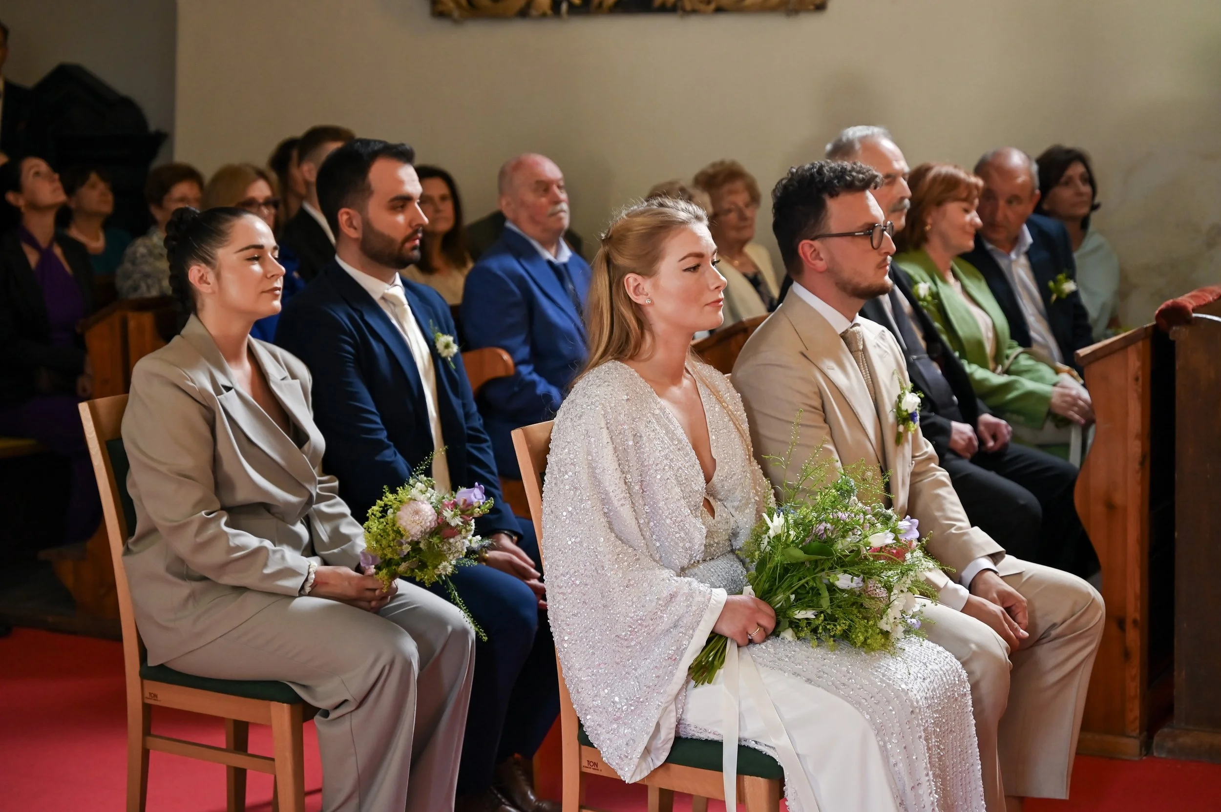 People attending a wedding ceremony sitting in a church pew, with the bride and groom in the front row holding bouquets of flowers.