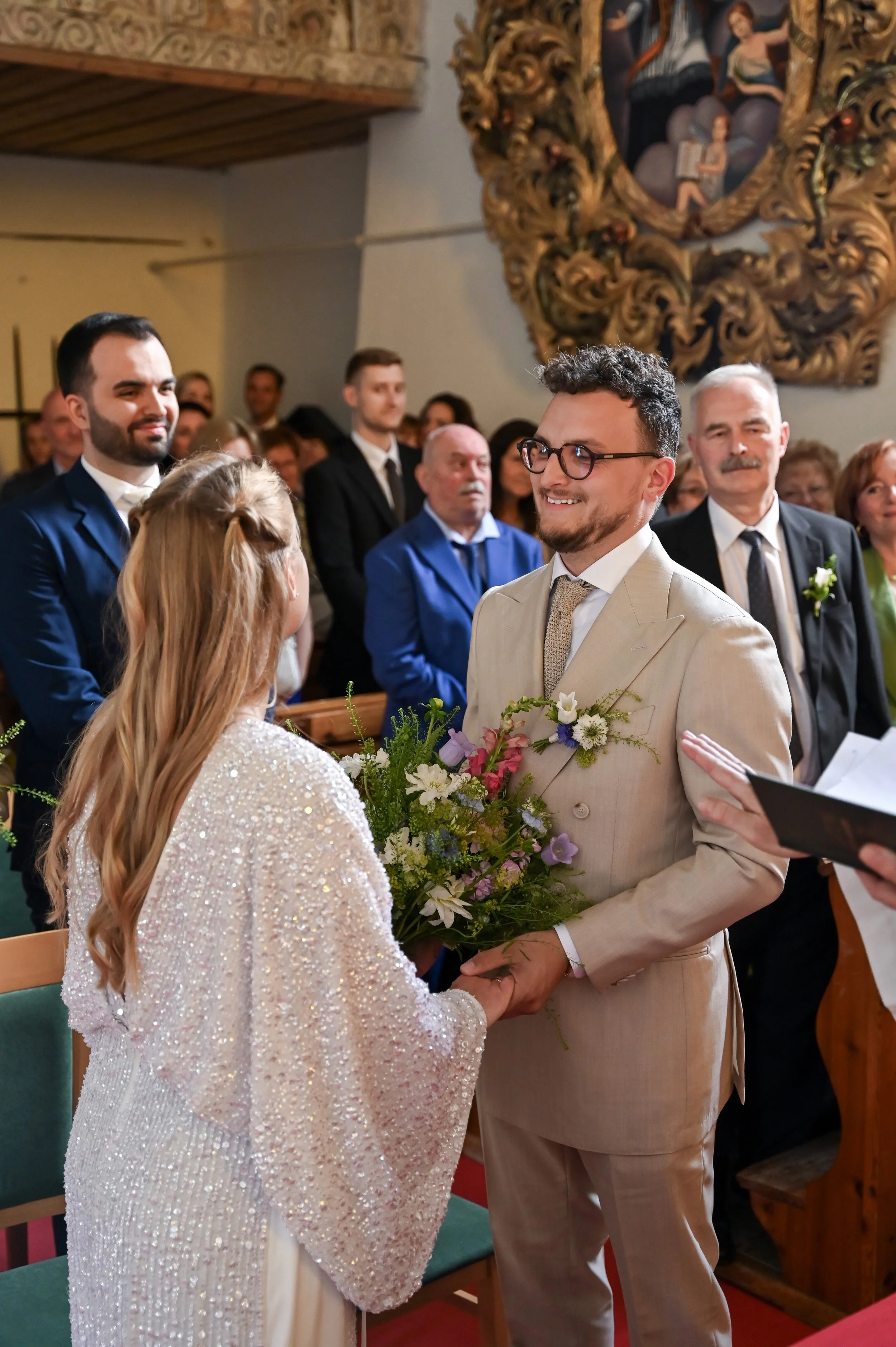 A man and woman exchanging vows and holding hands during a wedding ceremony inside a church, with guests seated behind them.