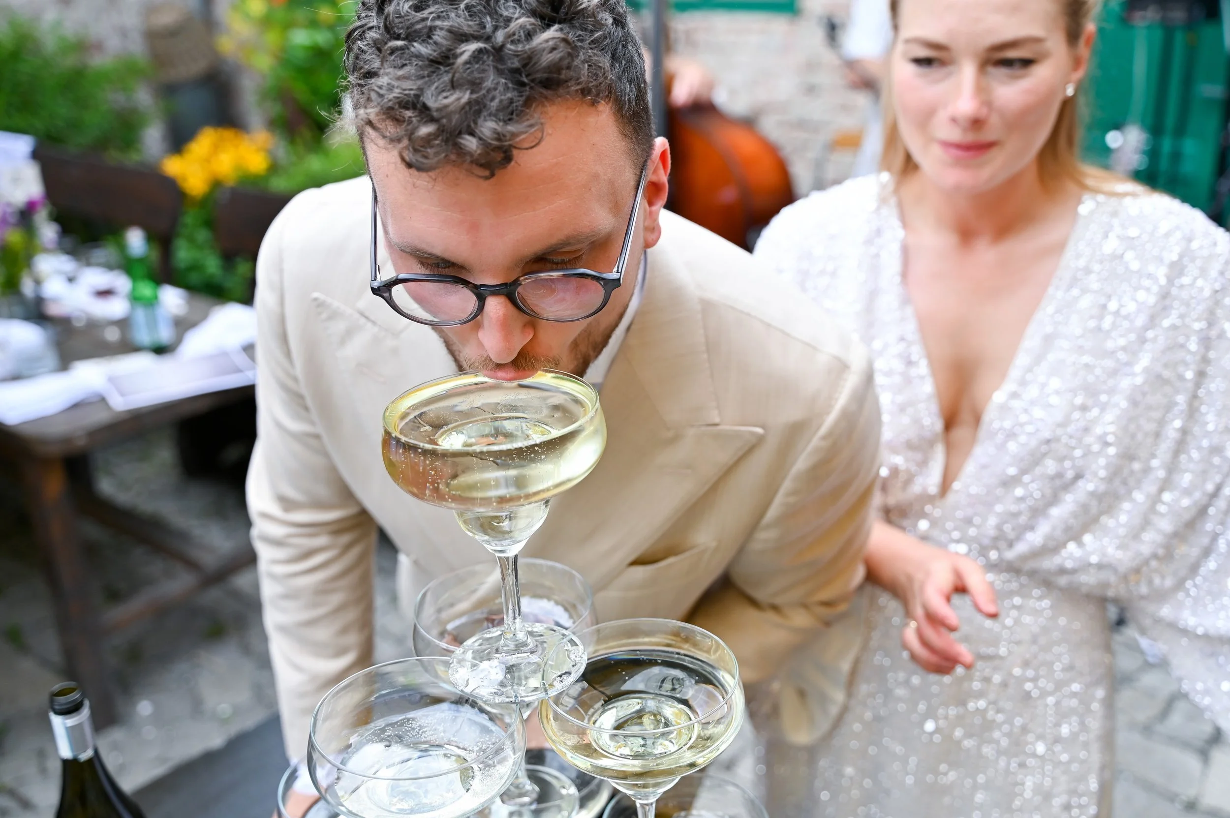 Man in a beige suit and glasses kissing the top of a champagne glass at a social gathering, with a woman in a sparkly dress watching nearby.