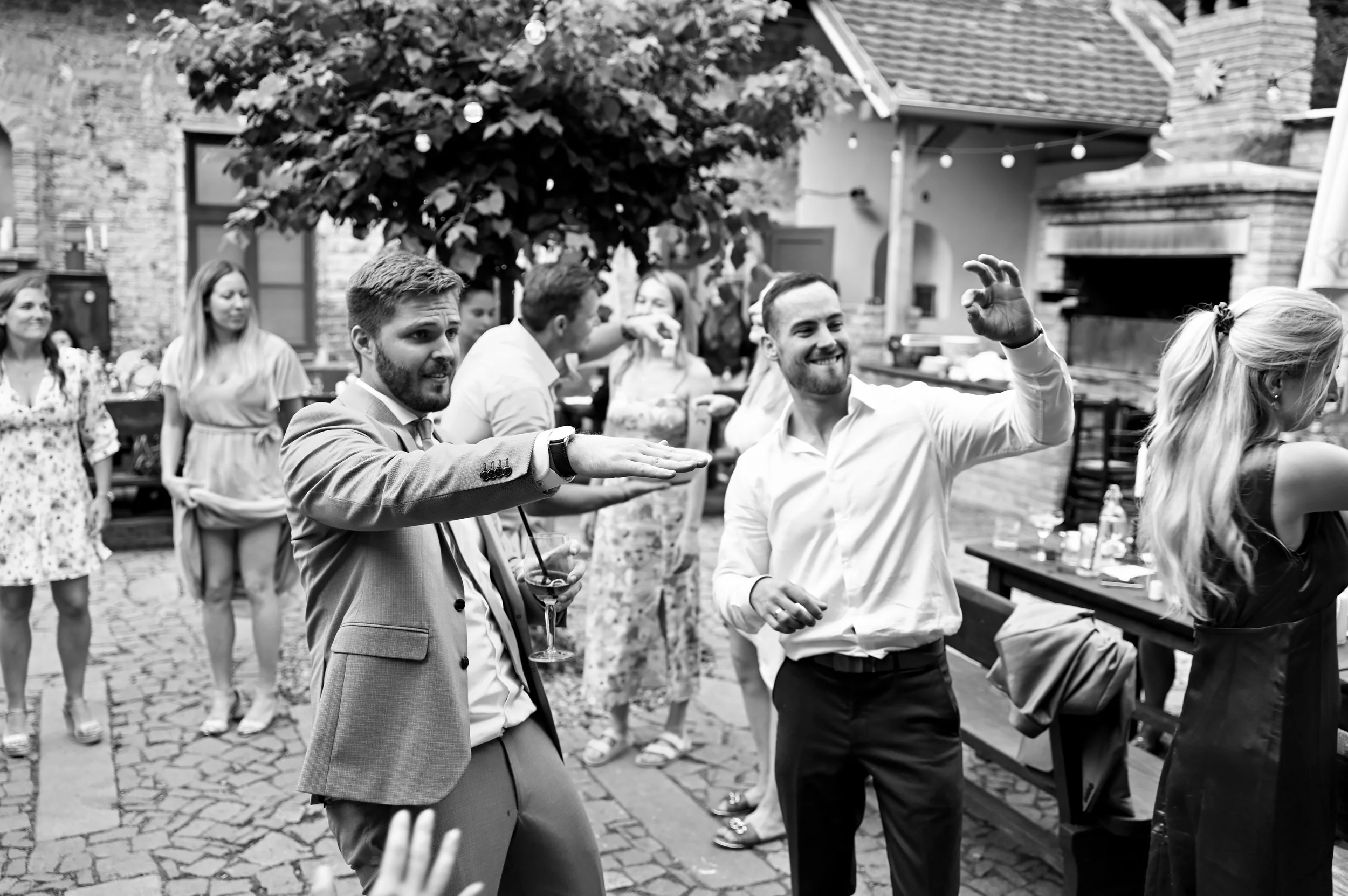 People dancing and socializing at an outdoor gathering or party, with a brick patio and string lights in the background.