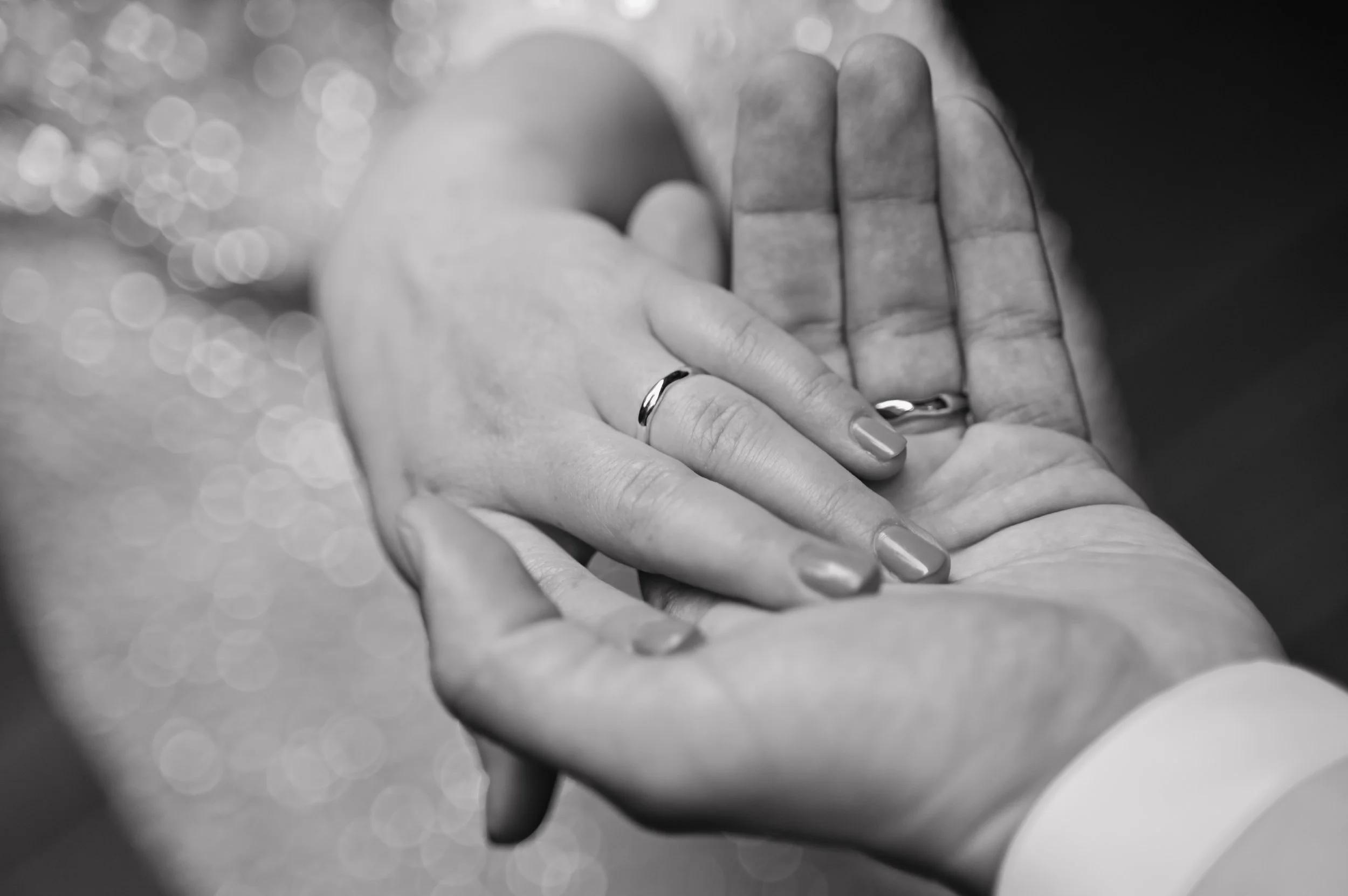 Two hands, one with a wedding band, gently holding each other. The image is in black and white with a blurred sparkling background.