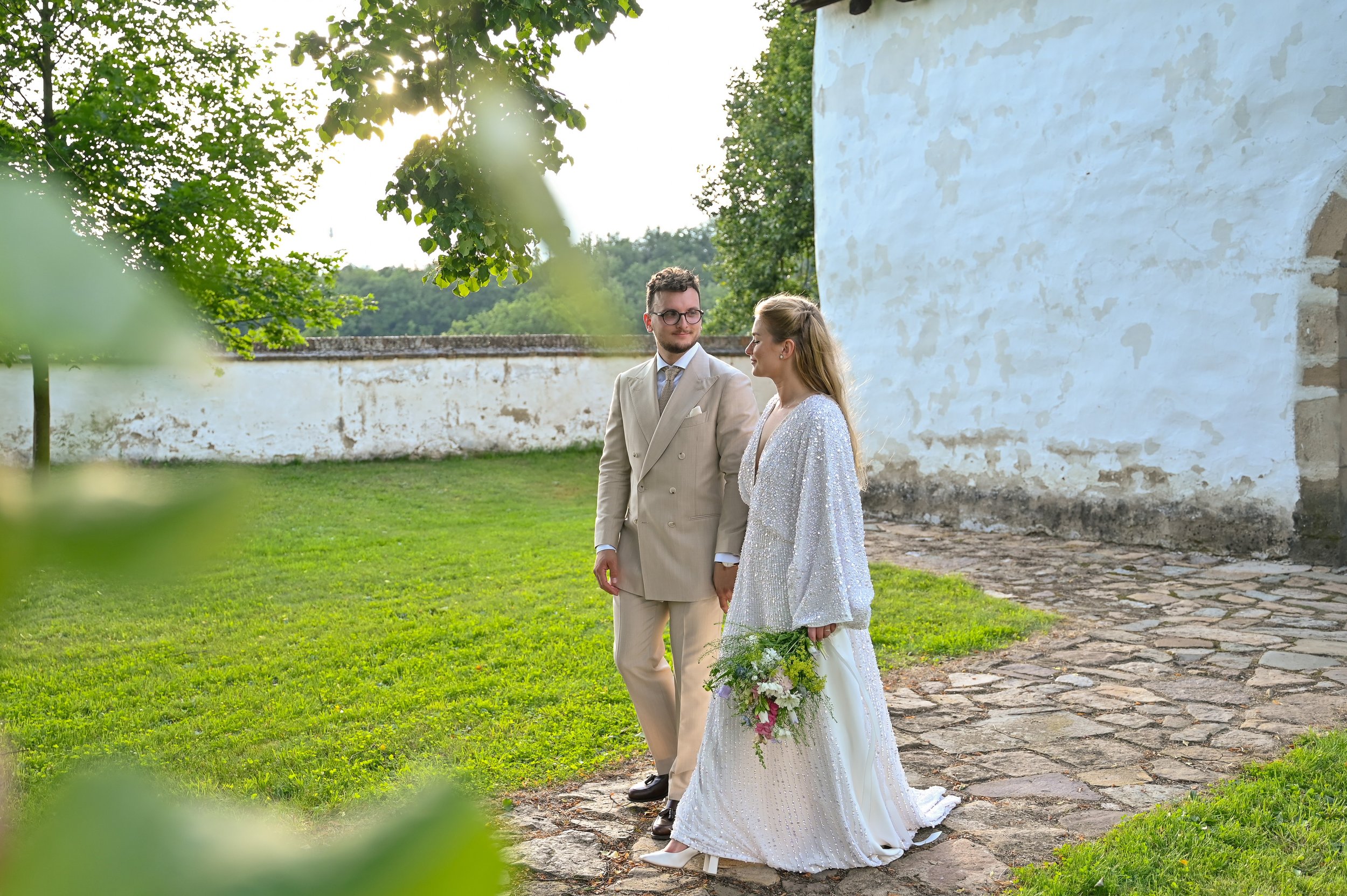 A bride and groom stand outdoors on a cobblestone path, holding hands, with lush green trees and a white weathered wall in the background, during daylight.