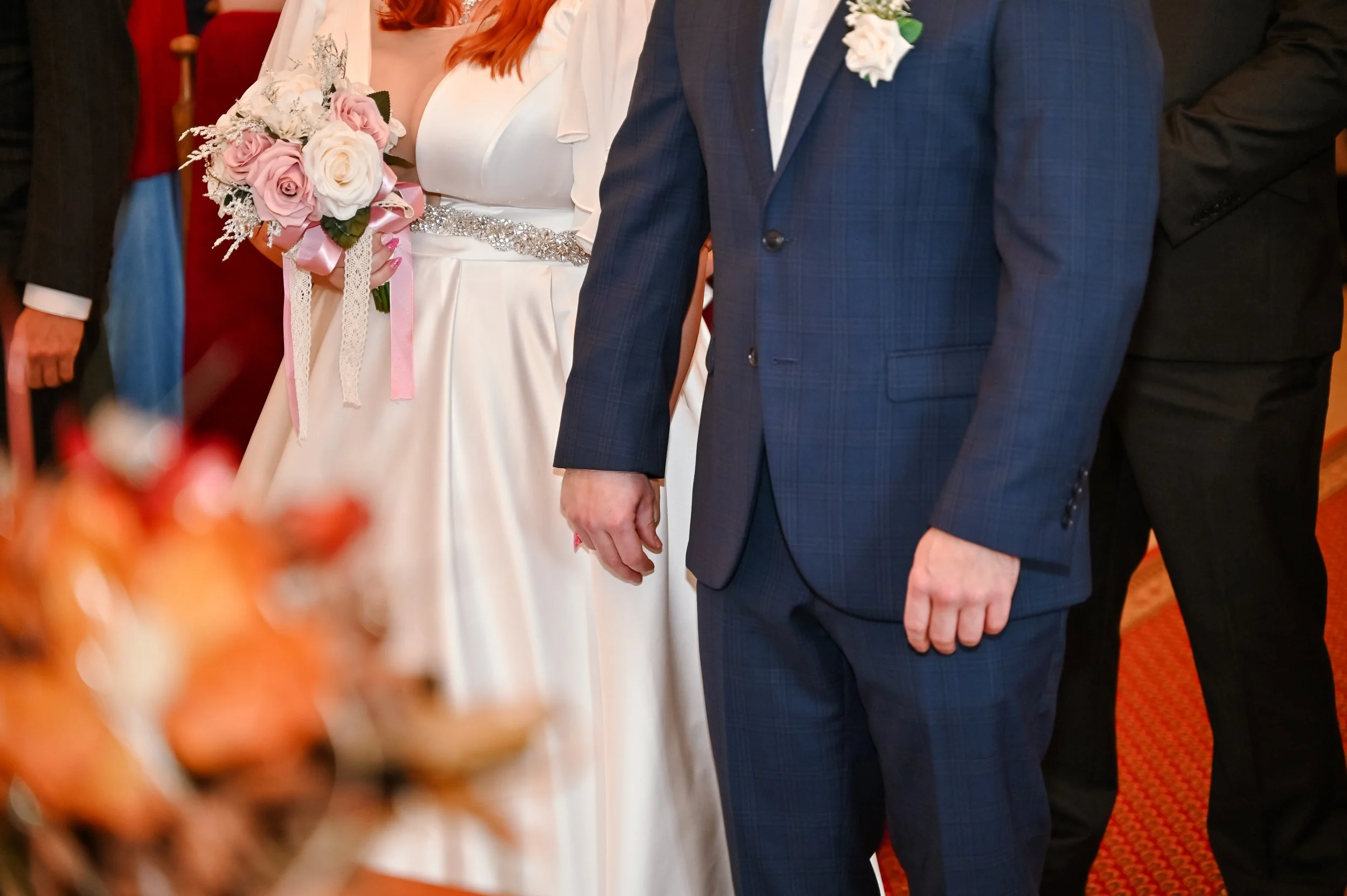 Close-up of a wedding ceremony showing a bride holding a bouquet of pink and white roses and a groom dressed in a blue plaid suit, with other guests in formal attire in the background.