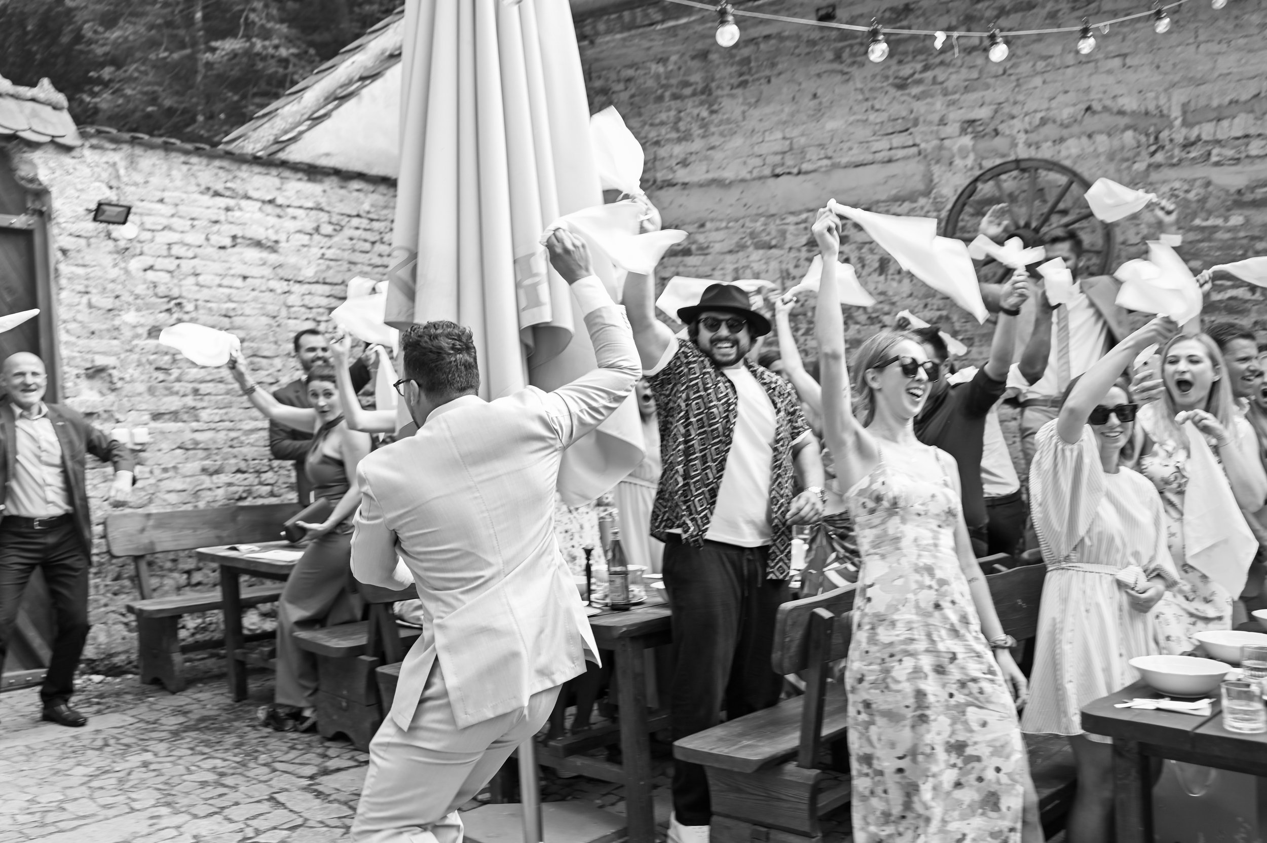People celebrating outdoors at a party, waving napkins in the air, smiling, and having fun on a cobblestone patio with brick walls and string lights.