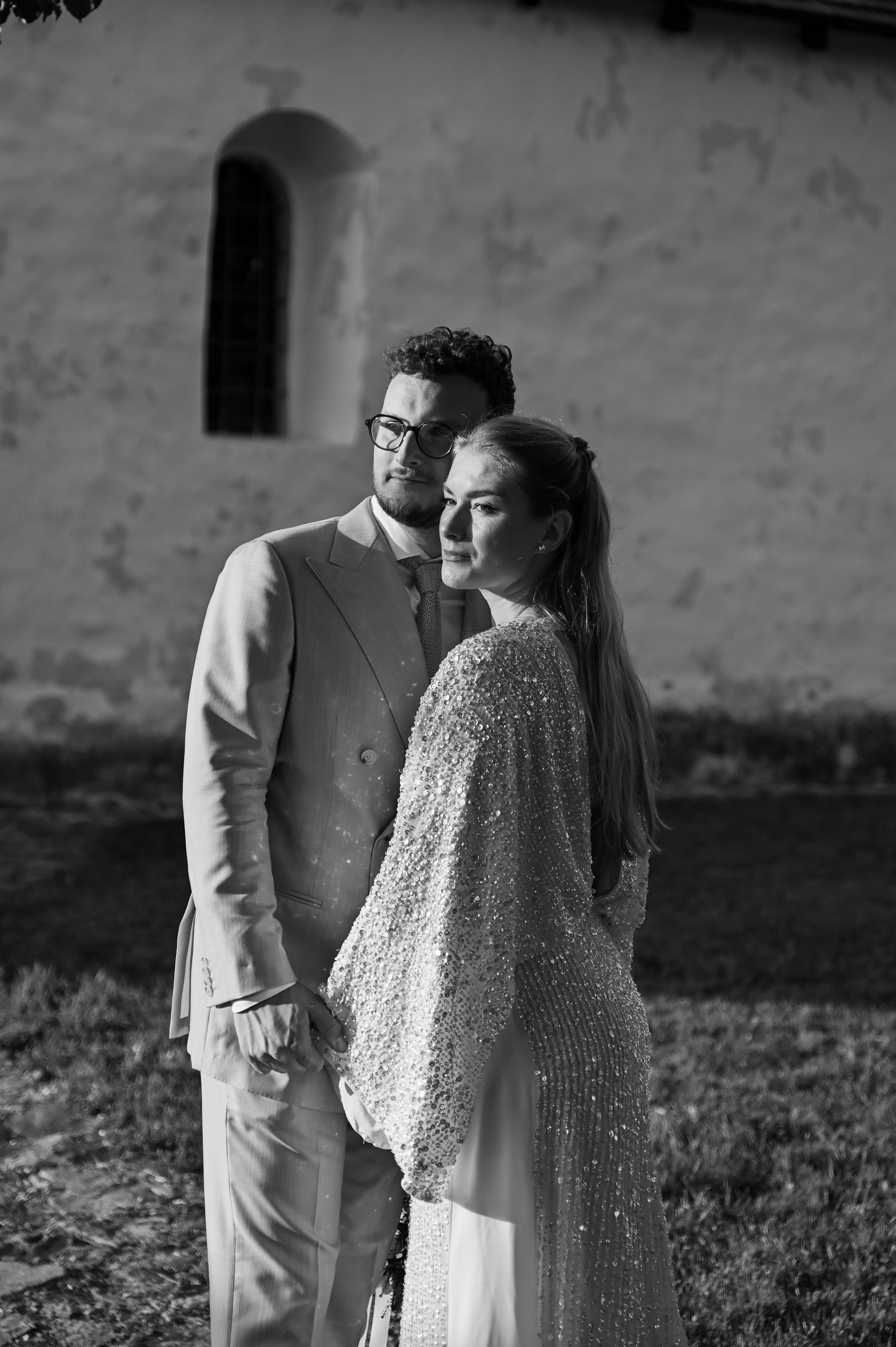 A black and white photo of a man and woman standing close together outdoors at night, holding hands, with a textured wall and narrow arched window in the background.
