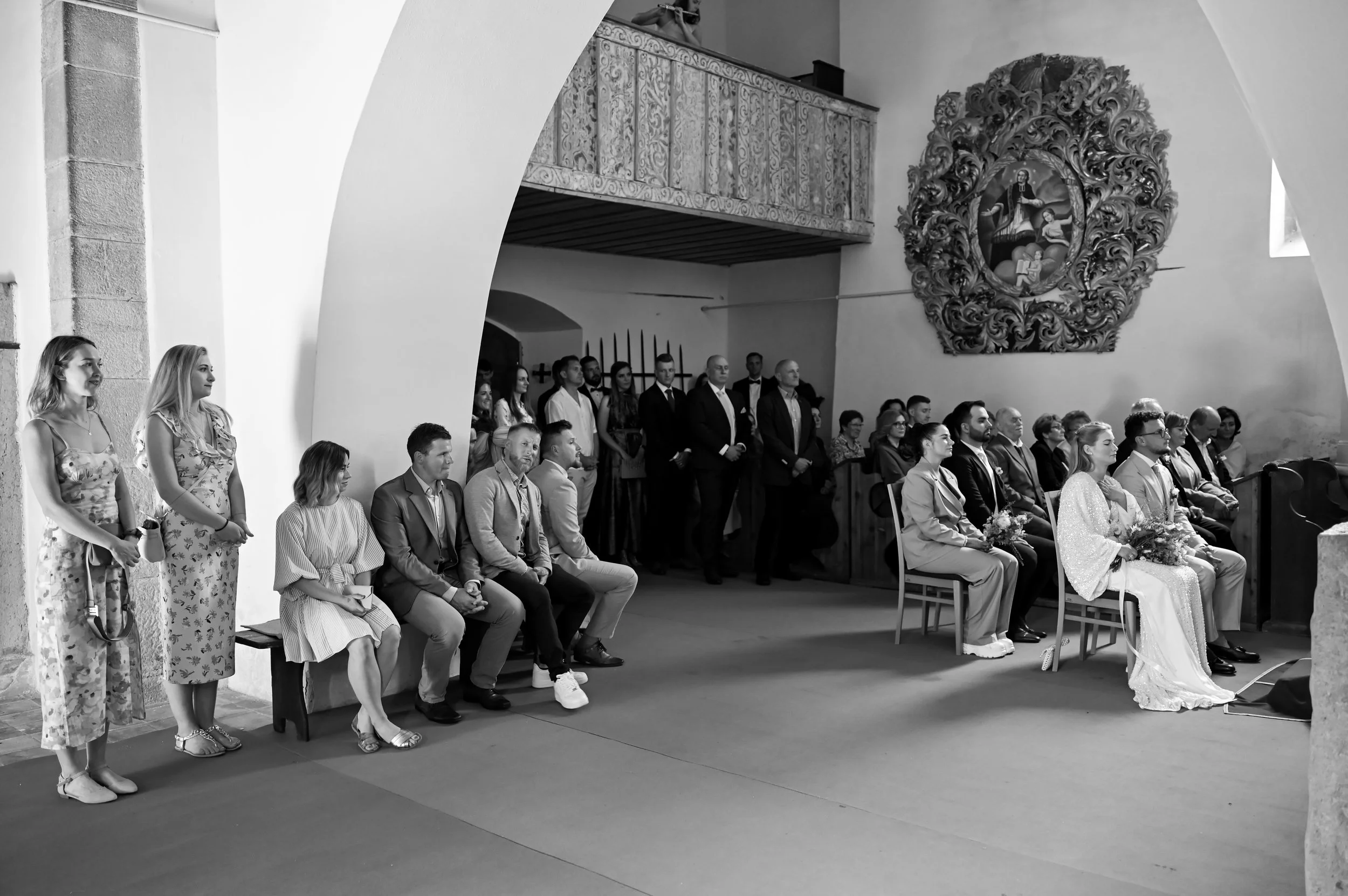 A black and white photo of a wedding ceremony inside a church. The bride and groom are sitting in front, holding hands and facing forward. Several guests are seated and standing around them, dressed in formal attire. There is a large religious artwor