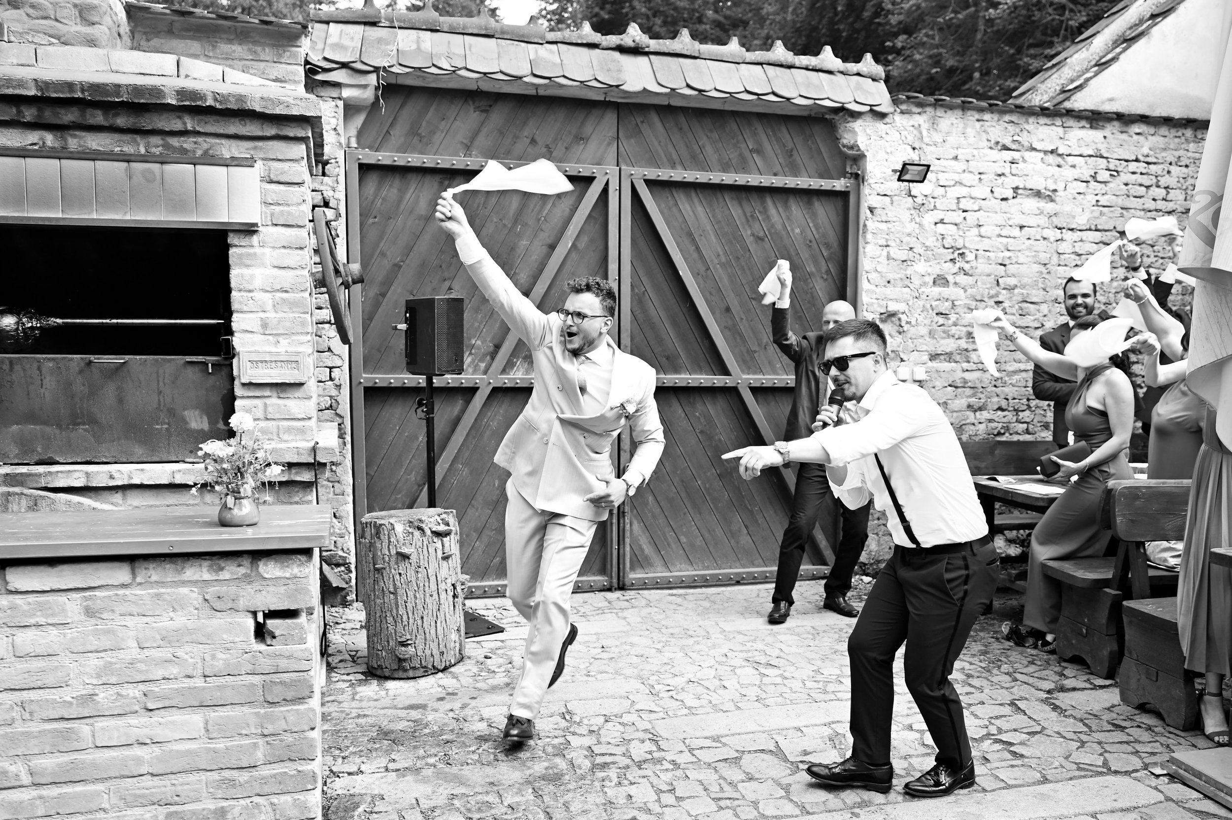 People celebrating at an outdoor event, dancing and waving napkins, with a man holding a microphone, on a cobblestone surface, with brick and wooden structures in the background.