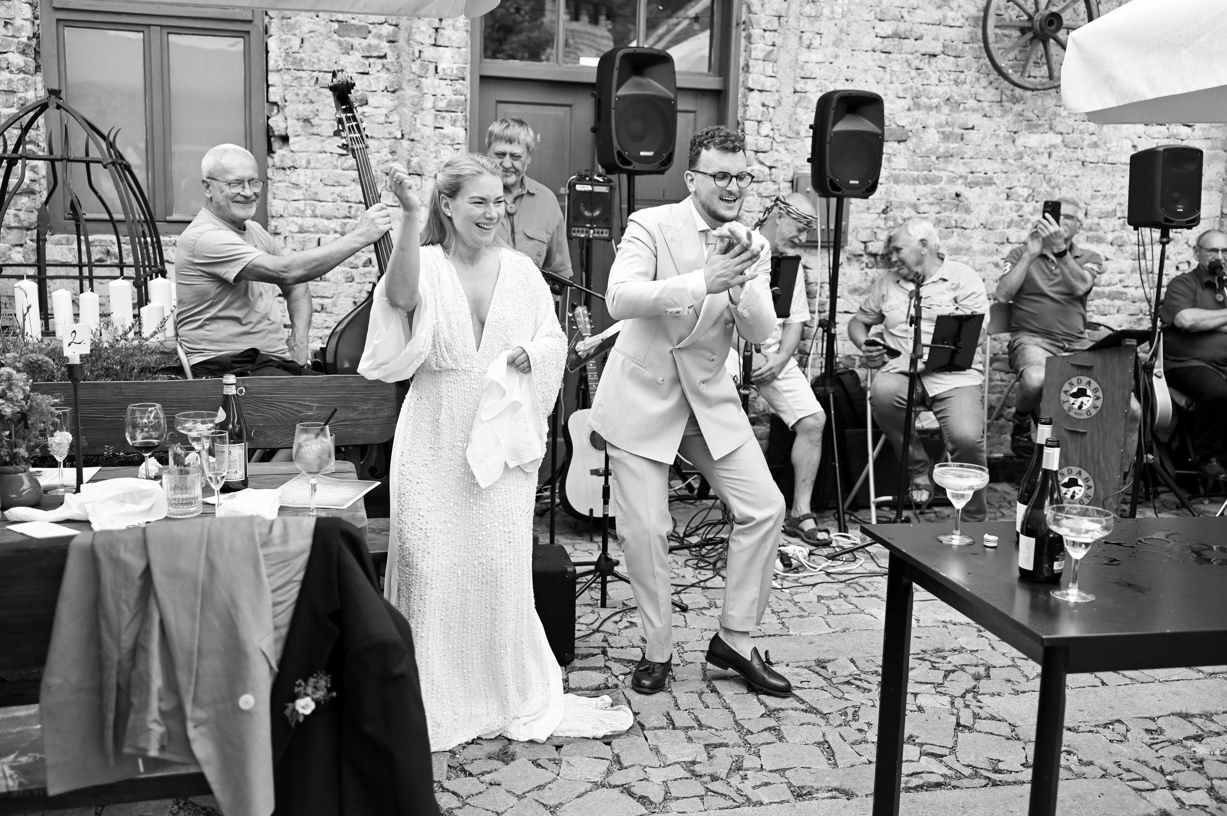 A black-and-white photo of a wedding reception outdoors with a brick wall background. The bride and groom are dancing, smiling, and holding hands. The bride is in a long dress holding a napkin, while the groom is in a suit. Around them, guests are se