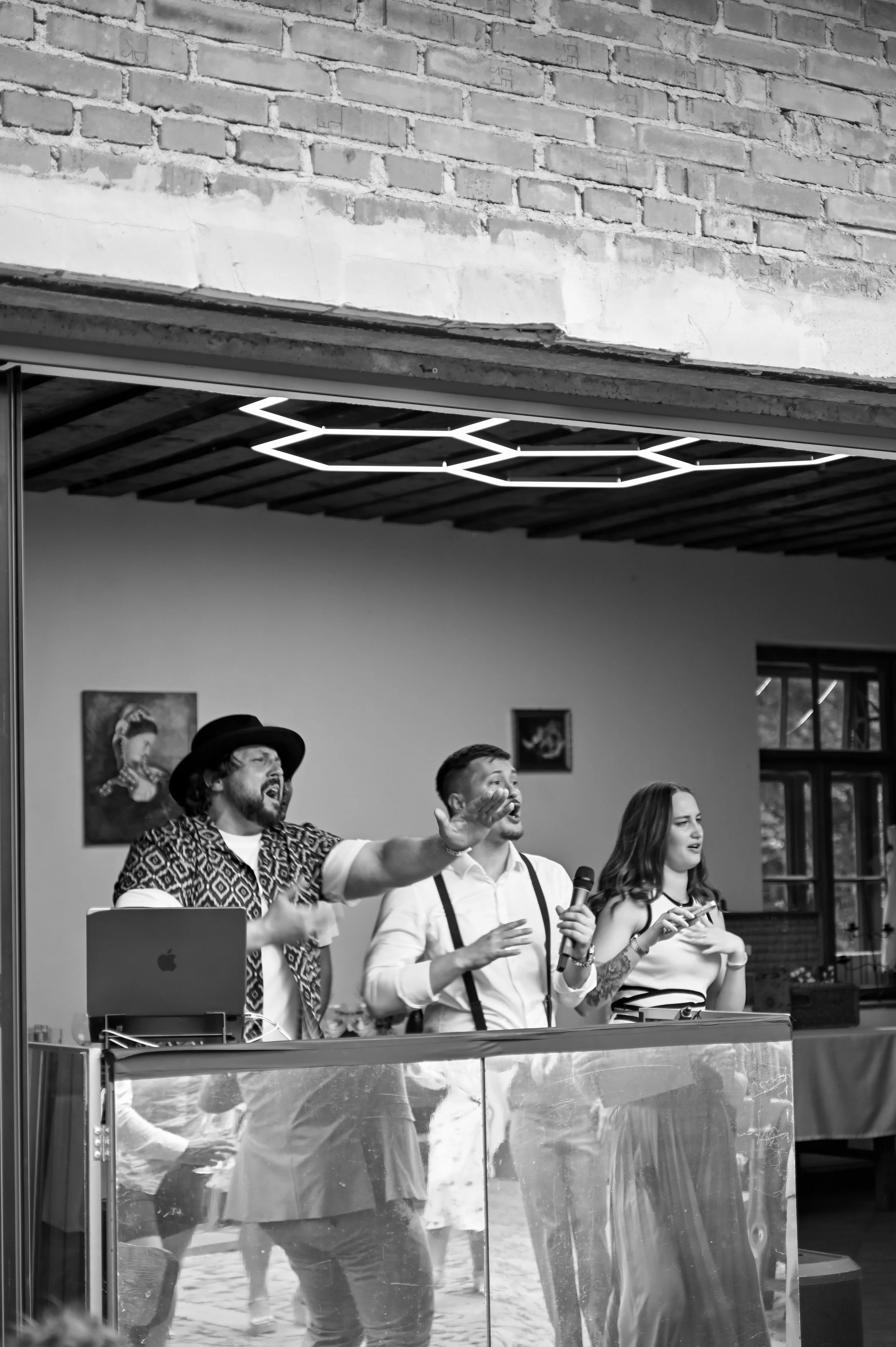 Three performers singing and speaking into microphones at an indoor event, with people dancing in the background. The scene is in black and white.