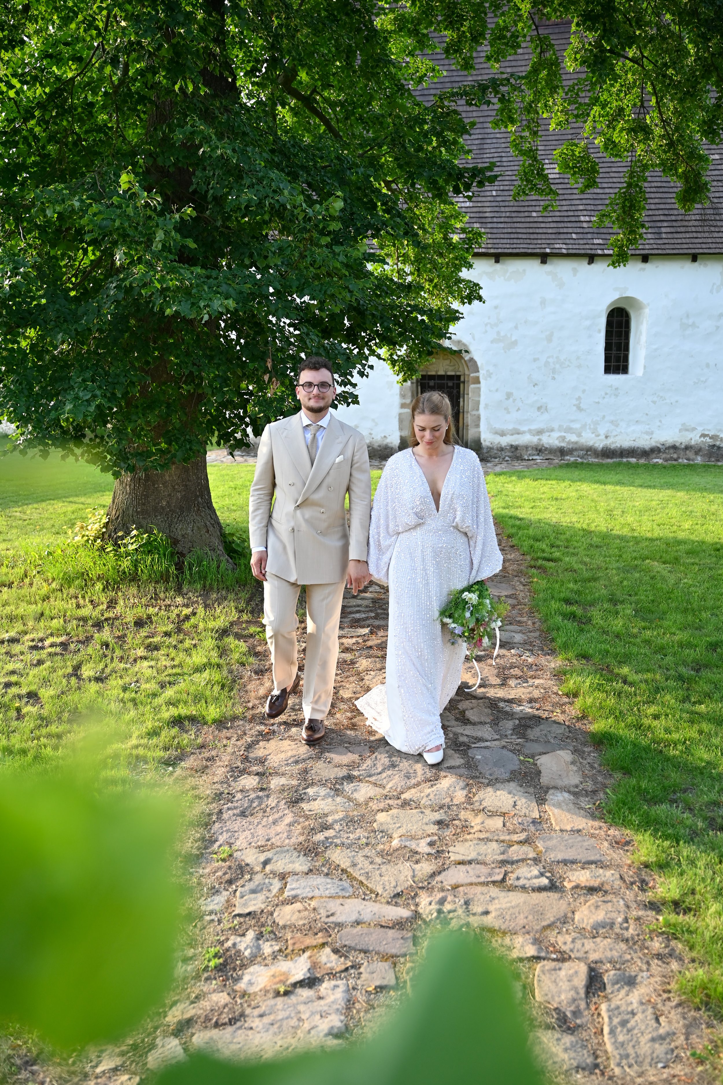 A newlywed couple walking hand-in-hand outdoors near a white church, with the bride in a white gown holding a bouquet and the groom in a beige suit, under a large leafy green tree.