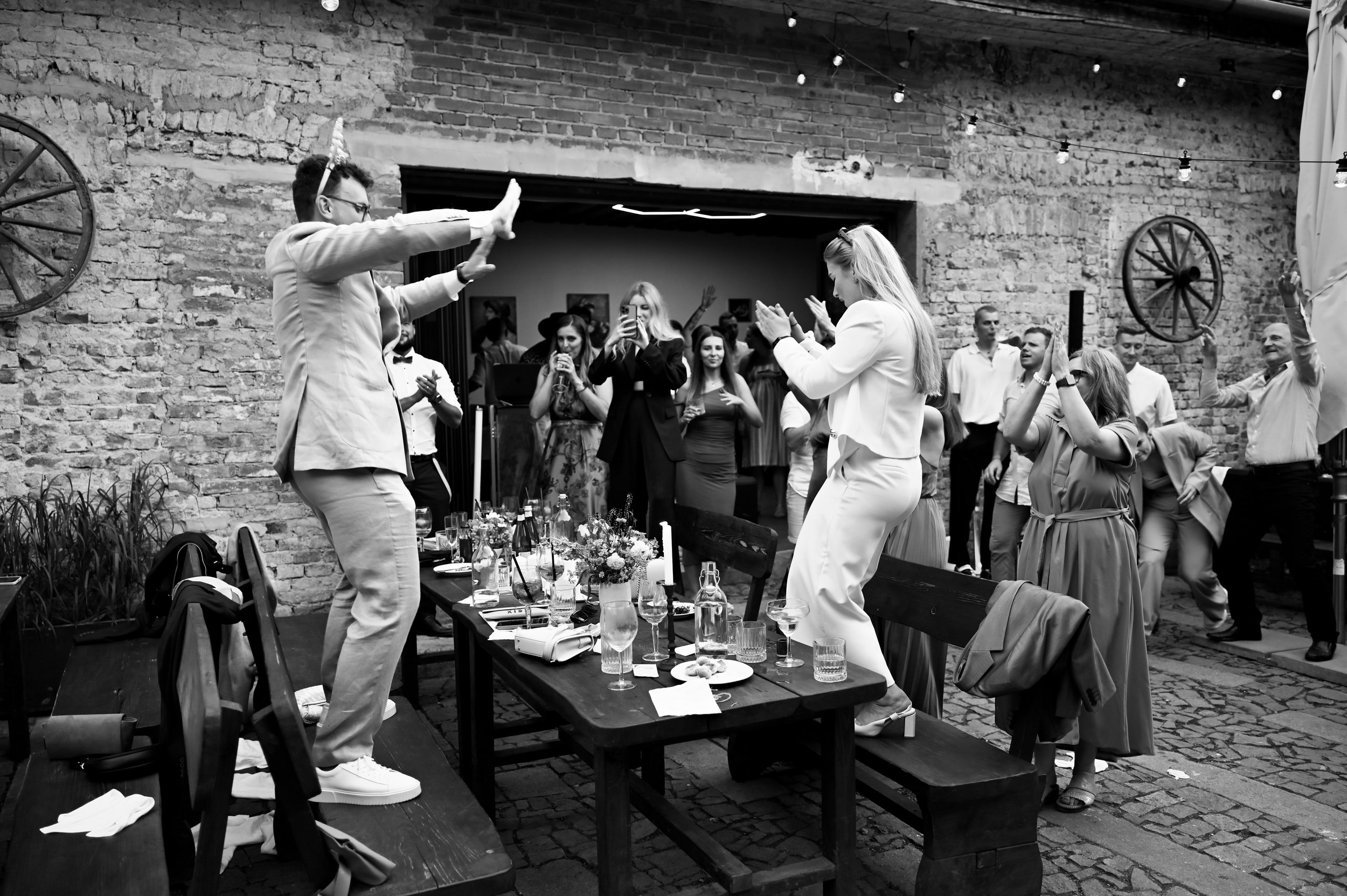 Guests dancing and celebrating at a lively party with a brick wall background and string lights.