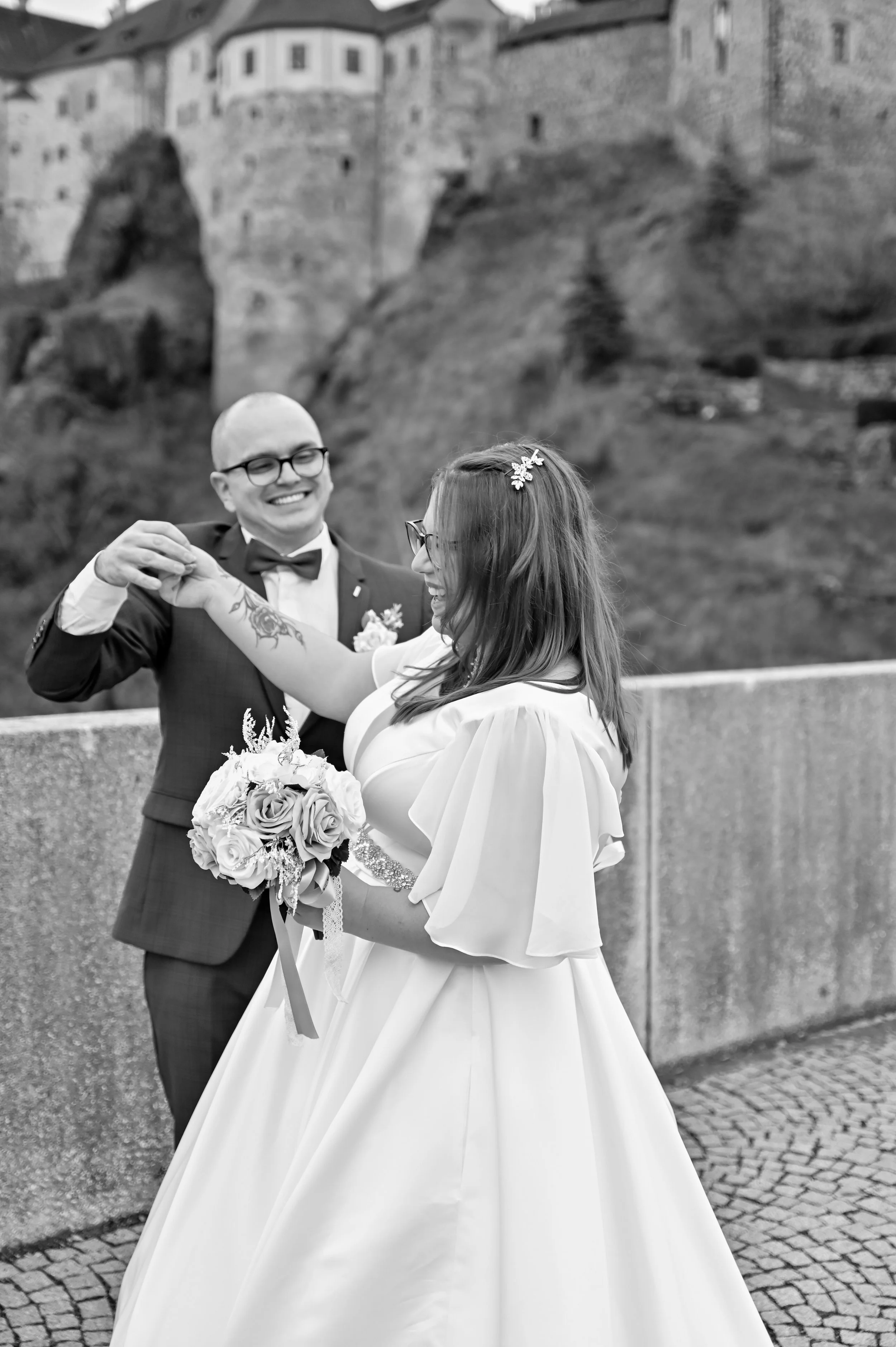 A newlywed couple dancing outdoors, with a castle on a hill in the background, black and white photo.