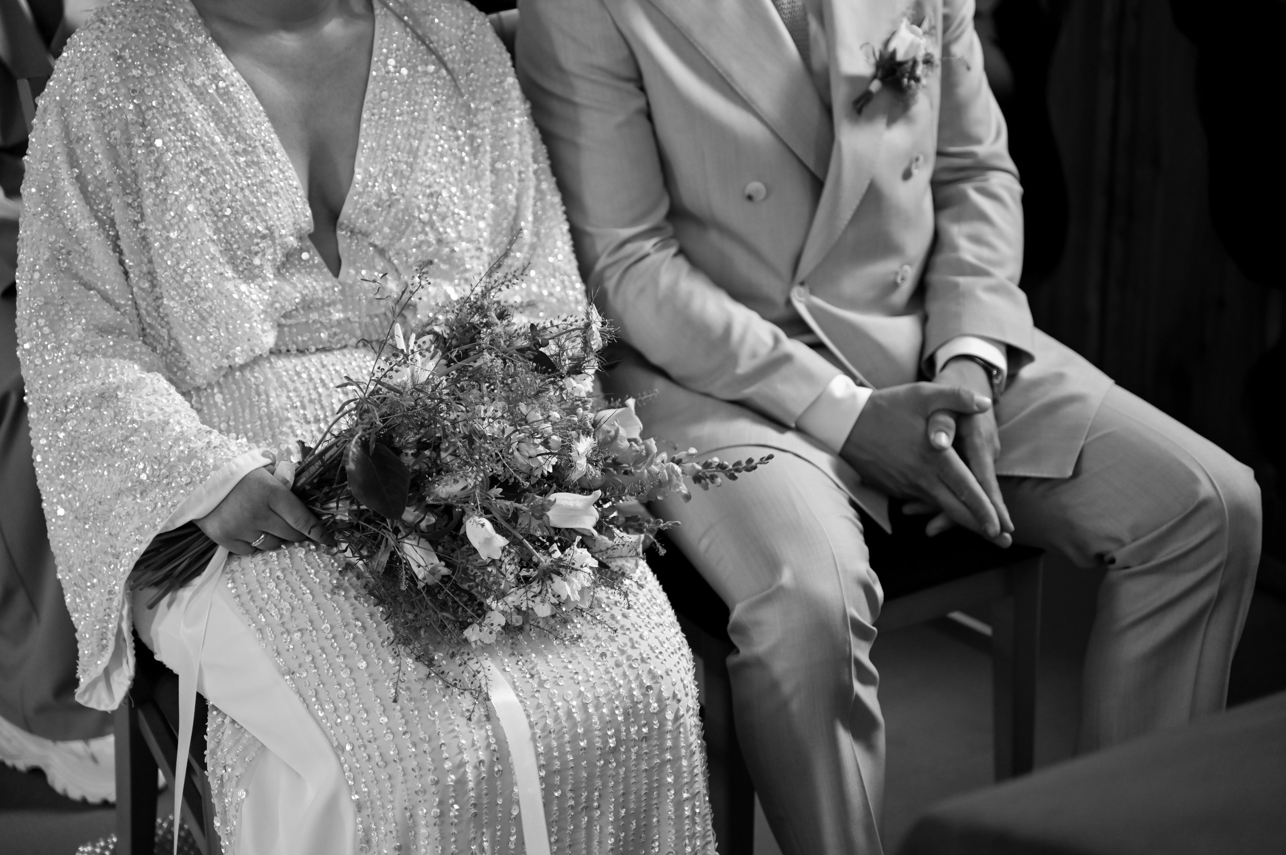 Close-up of a bride holding a bouquet of flowers and a groom sitting with hands clasped, during a wedding ceremony in black and white.