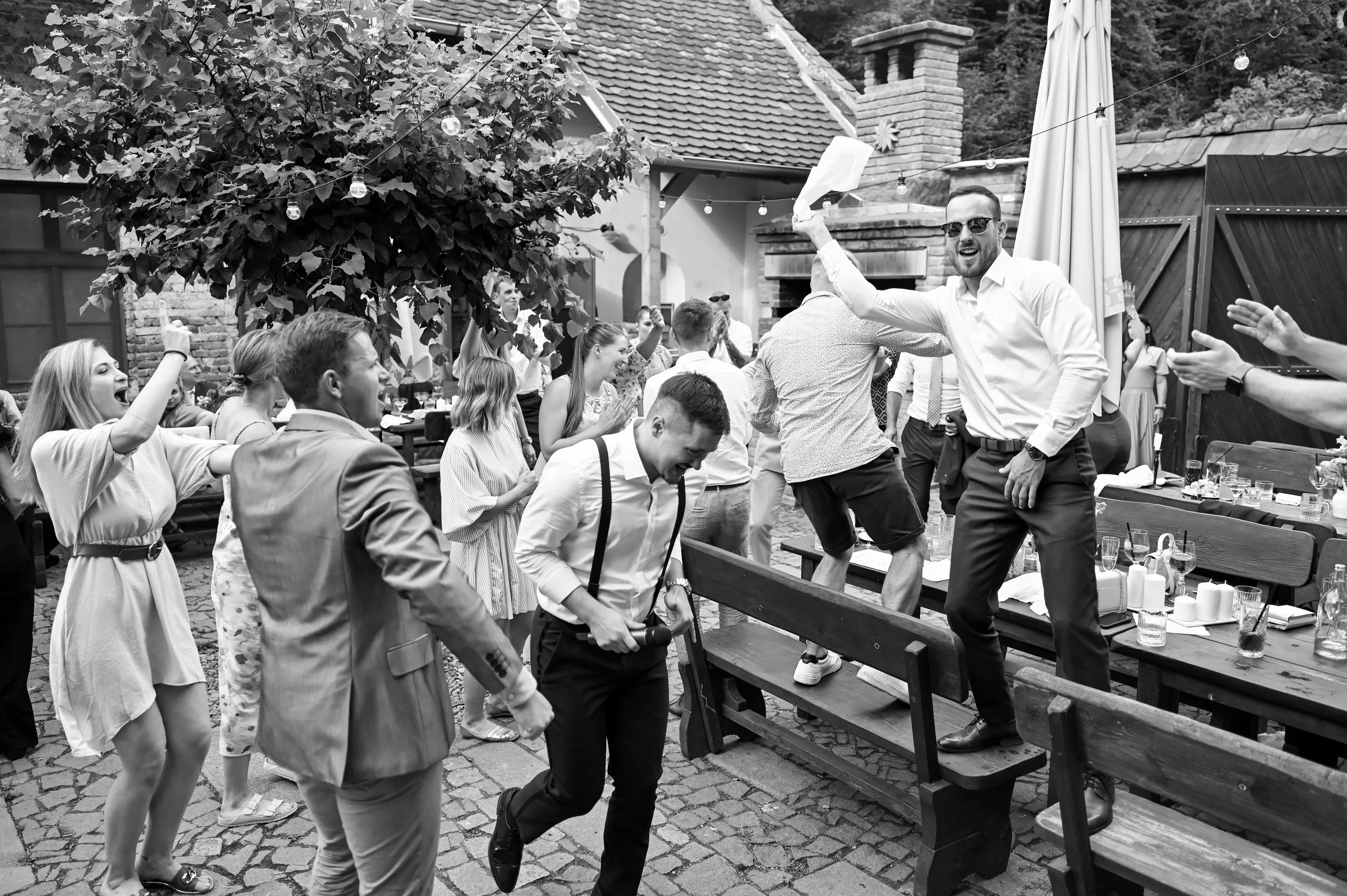 People at a lively outdoor party dancing and celebrating, with some on benches and others standing and enjoying the event in a backyard setting.