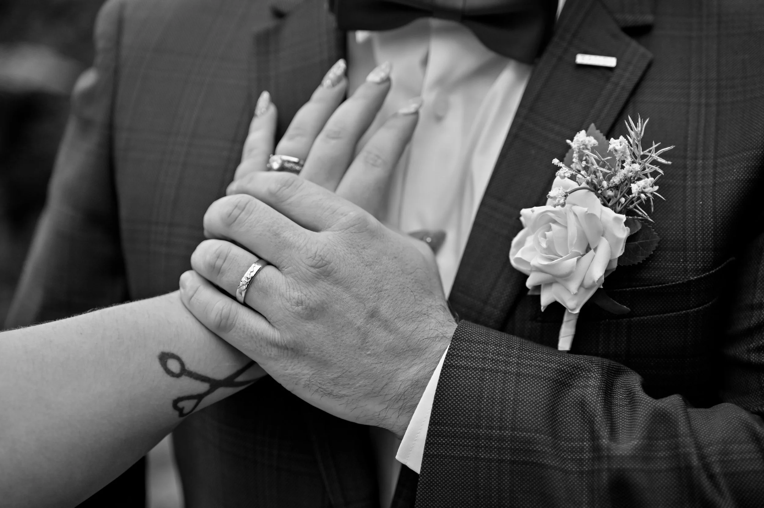 Close-up black-and-white photo of a groom and bride holding hands, showing wedding rings. The groom is wearing a suit with a boutonnière, and the bride has a tattoo of scissors on her wrist.