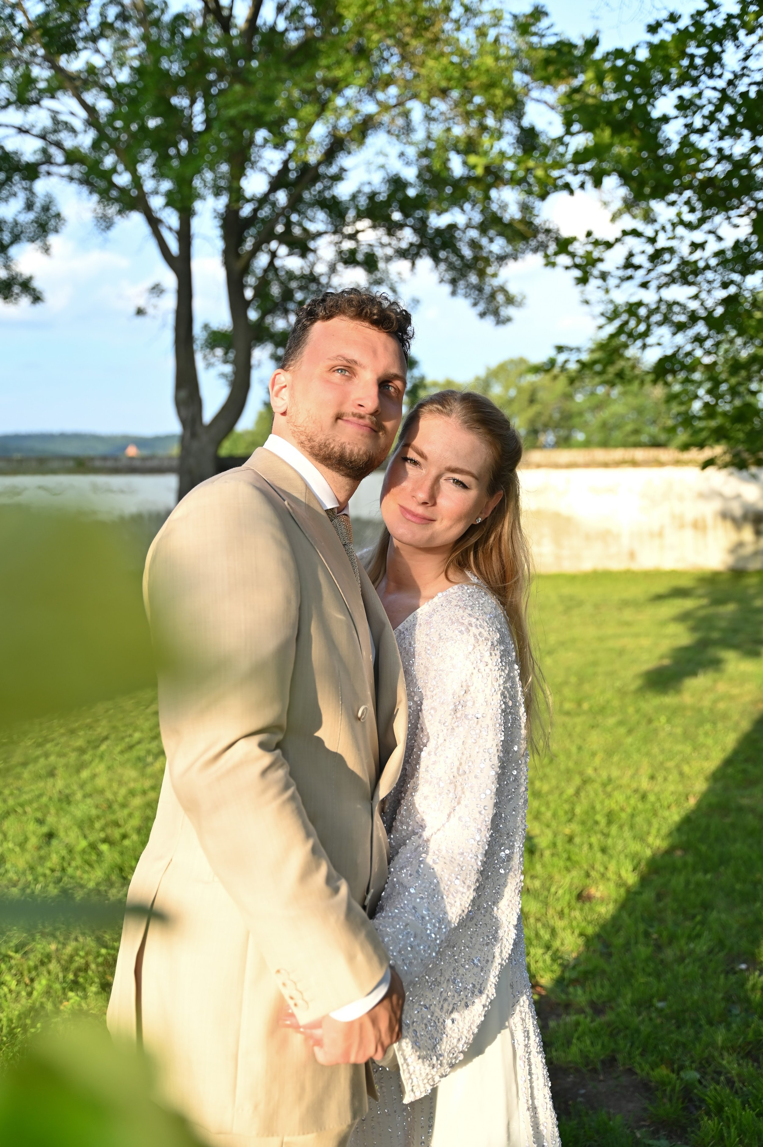 A newlywed couple posing outdoors on a sunny day with trees and a stone wall in the background.