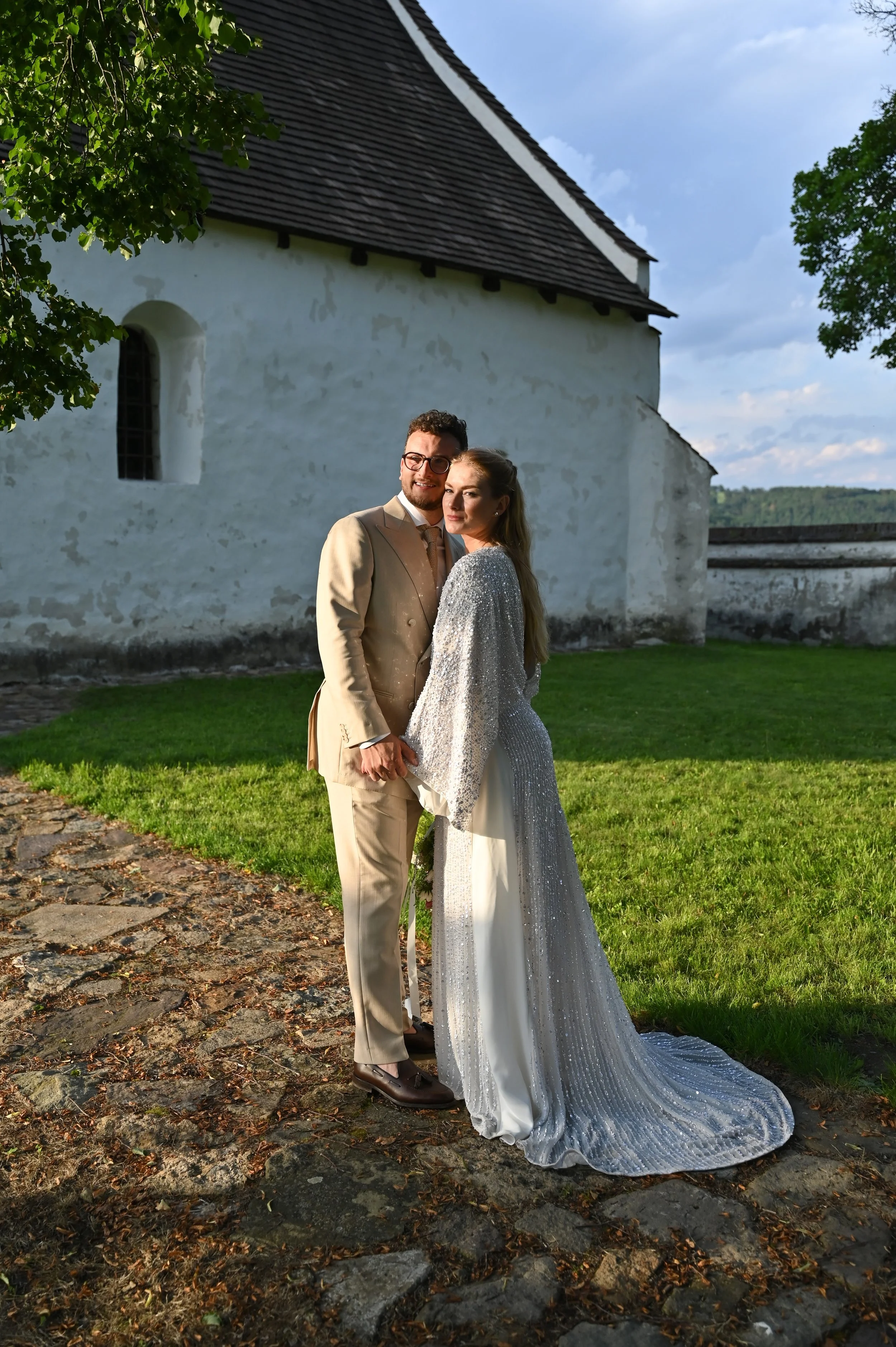 A bride and groom standing outdoors on a stone pathway near a white church with a dark roof, during late afternoon or early evening. The bride is wearing a glittering silver gown with wide sleeves, and the groom is dressed in a beige suit with brown 