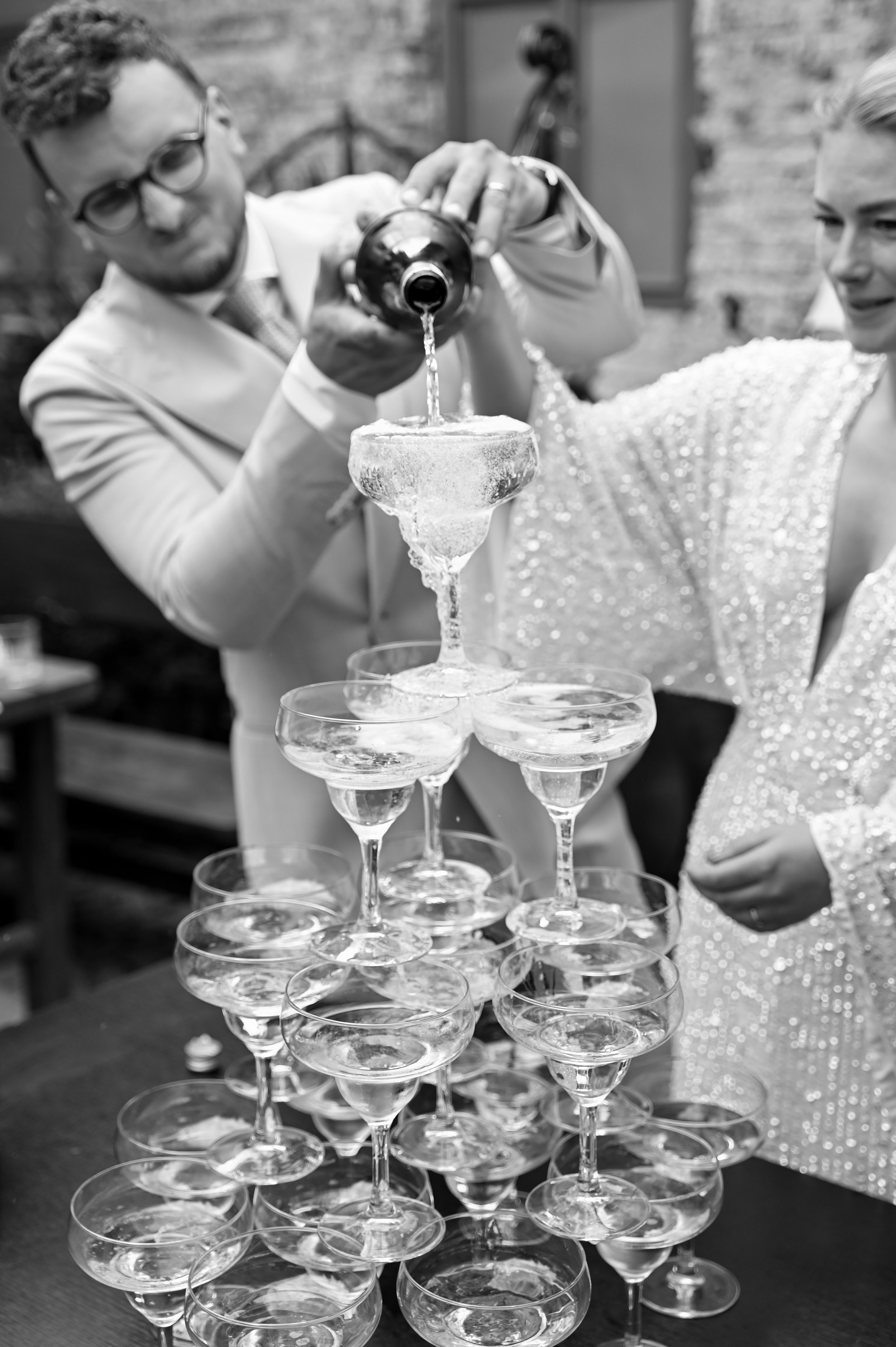 Two people, dressed in elegant attire, are pouring champagne into a pyramid of coupe glasses at a celebration or event.