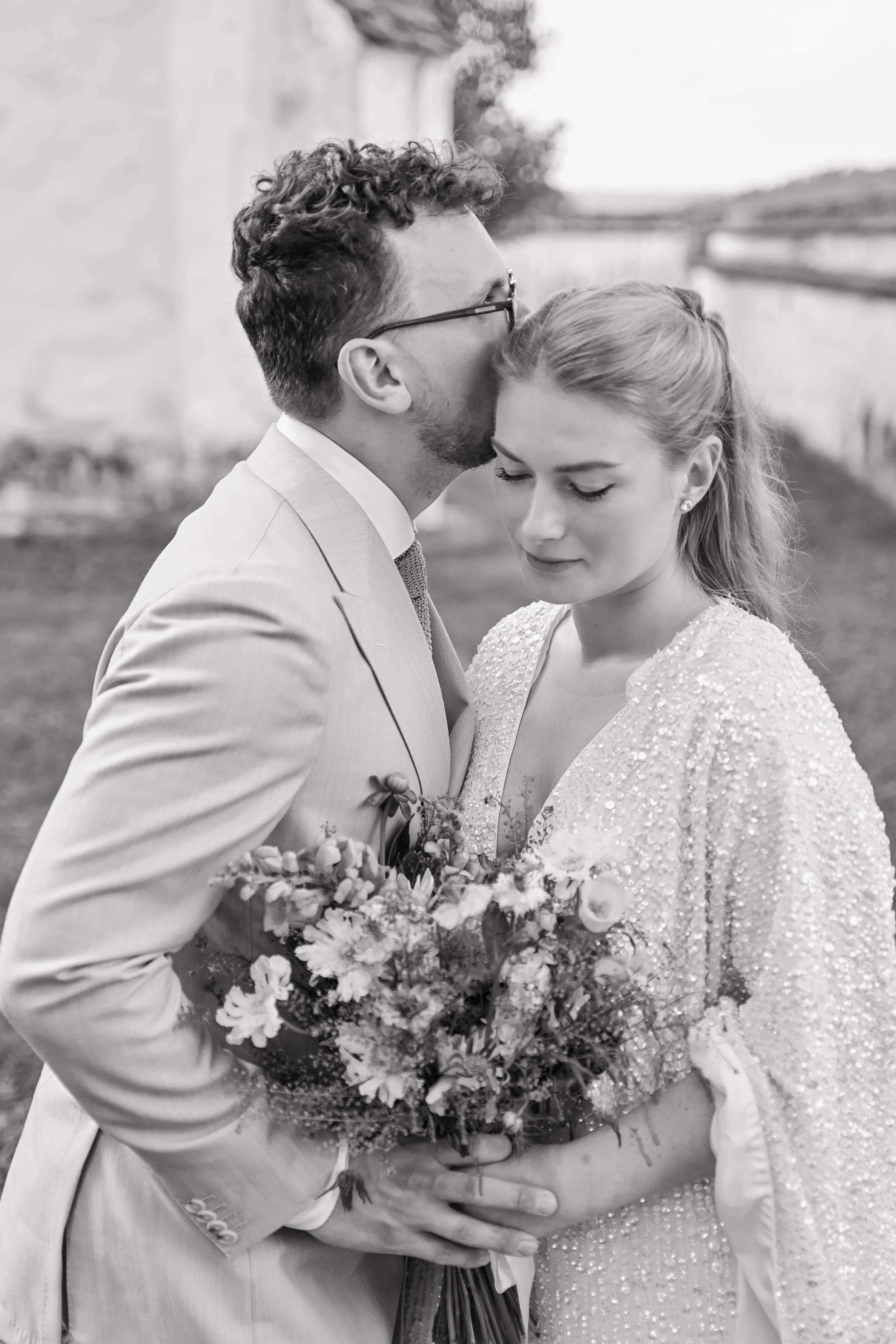 A black and white photo of a wedding couple, the groom kissing the bride on the forehead while she holds a bouquet of flowers with her eyes closed.