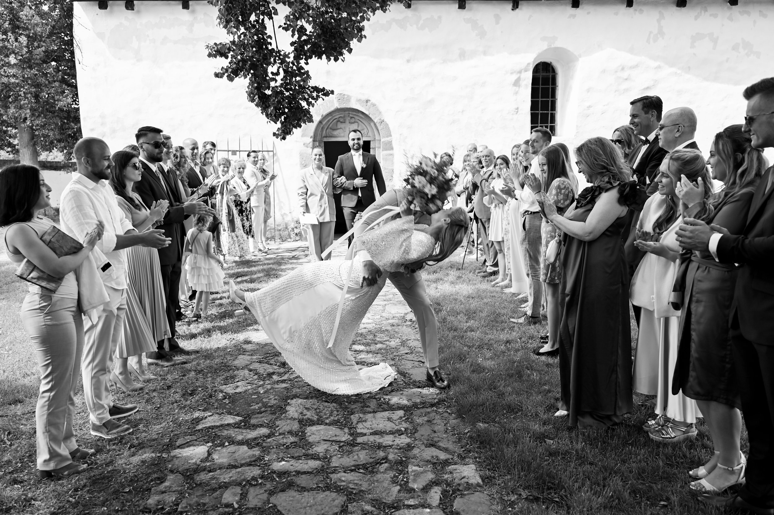 A bride tipping upside down during a wedding ceremony outdoors, surrounded by guests clapping and smiling in front of a white church building.