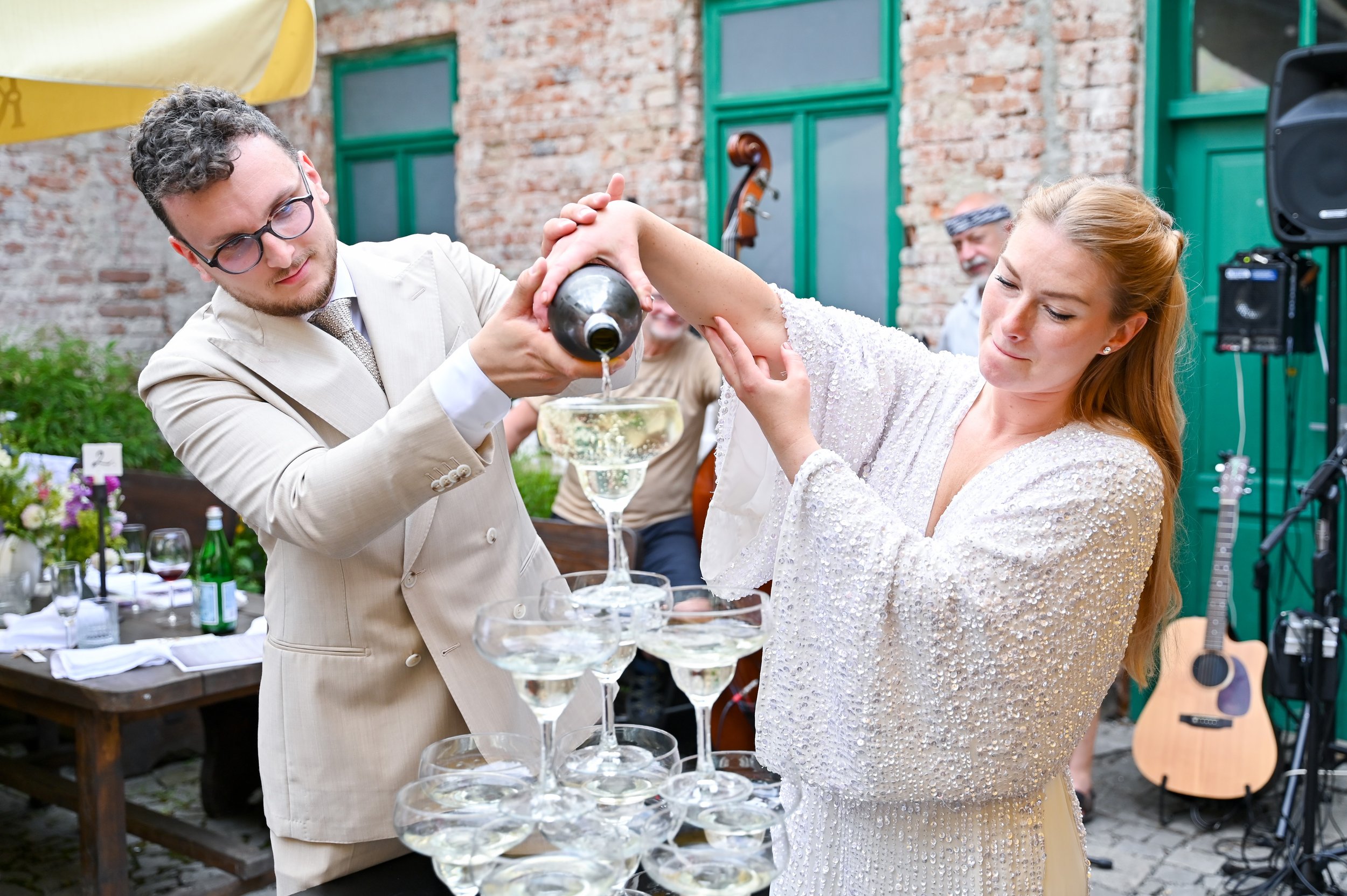 A woman in a white dress drinks champagne poured by a man in a beige suit at an outdoor celebration, with a table of glasses and a guitar in the background.