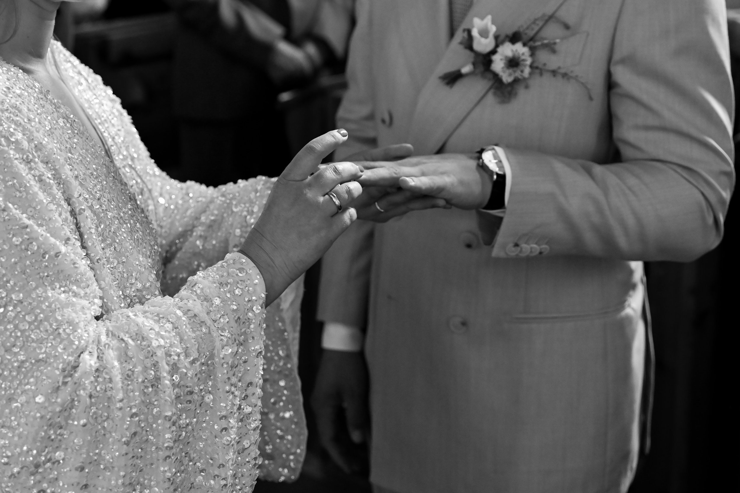 A bride and groom exchanging rings during a wedding ceremony, with the groom placing a ring on the bride's finger, in black and white.