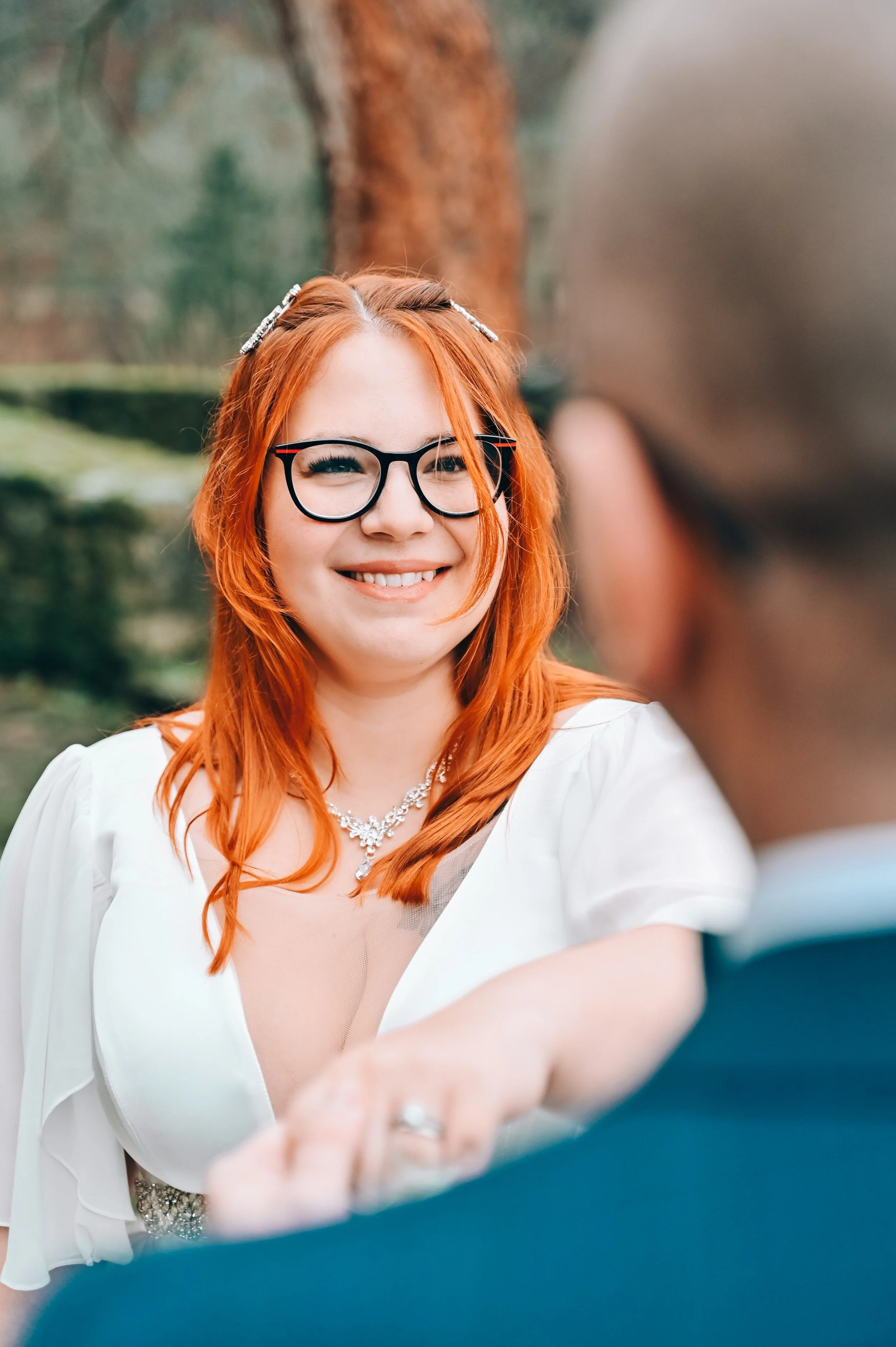 Woman with red hair, wearing glasses, a white dress with jewelry, smiling at a man outdoors with trees in the background.