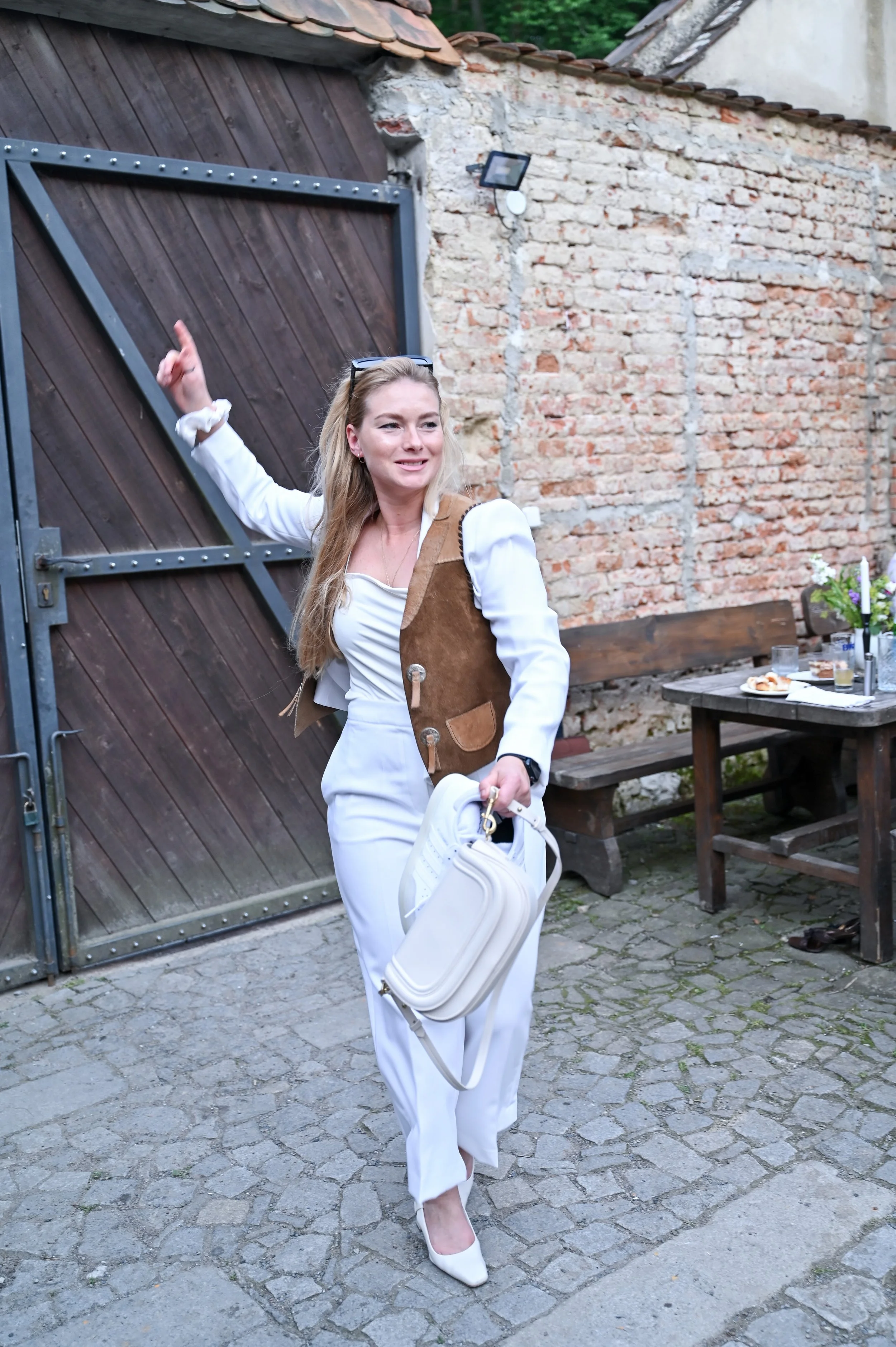 A woman dressed in white with a brown vest, holding a white handbag, pointing upward and smiling outdoors in front of a brick and wood wall with a wooden table and bench nearby.