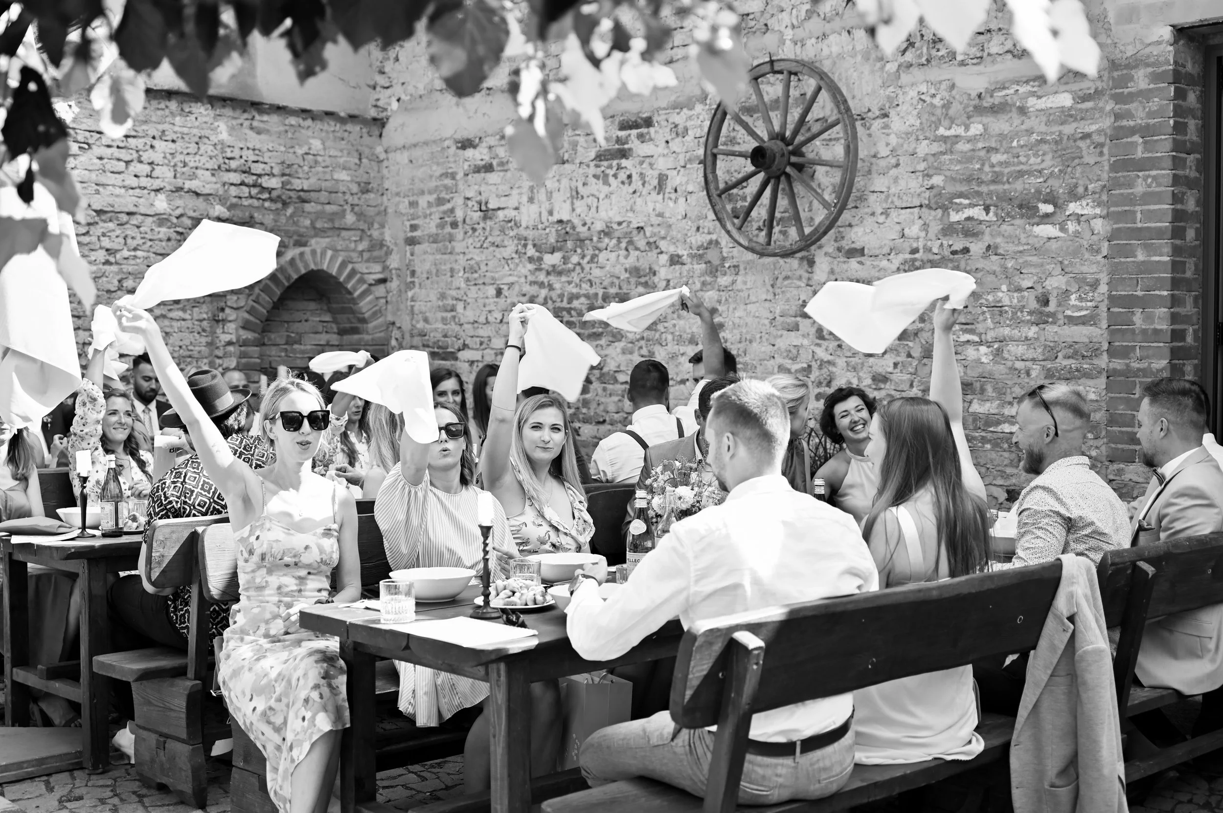 A black and white photo of a group of people sitting at an outdoor table, enjoying a meal and waving napkins in the air. They are in a courtyard with a brick wall and a wooden wheel hanging on the wall.