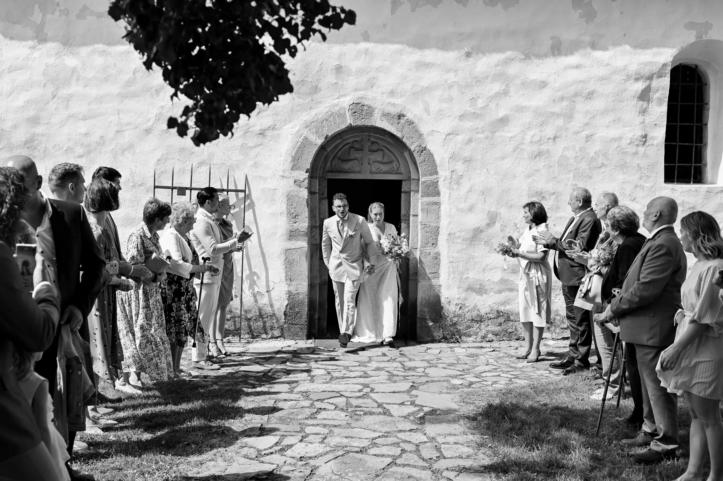 Black and white photo of a wedding ceremony outside a stone building, with the bride and groom exiting and guests clapping and taking photos.