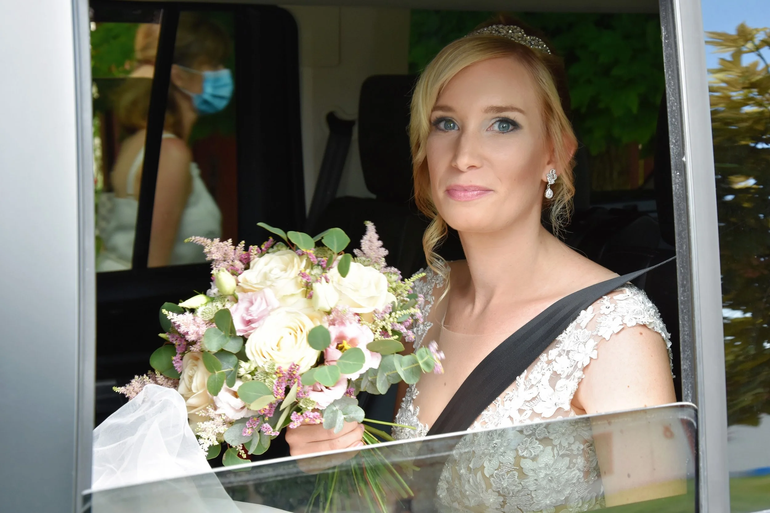 Nevěsta s blond vlasy, oblečená v krajkových svatebních šatech a špercích, sedící ve vozidle s kyticí květin, s osobou v masce viditelnou v pozadí z okna.