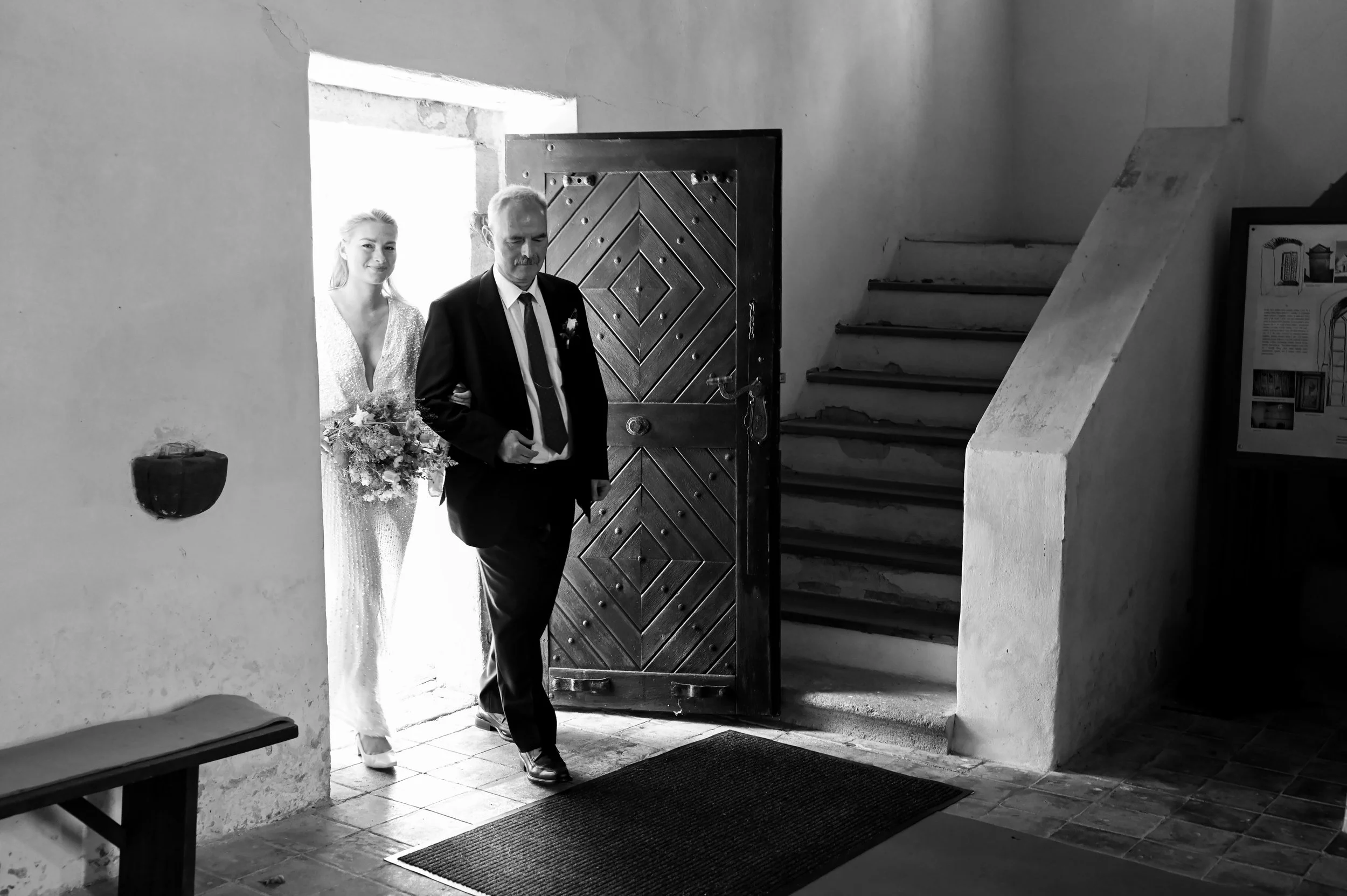 A man in a suit and a woman in a white dress with a bouquet of flowers are standing at the entrance of a building. The man appears to be walking out, while the woman is standing behind him.