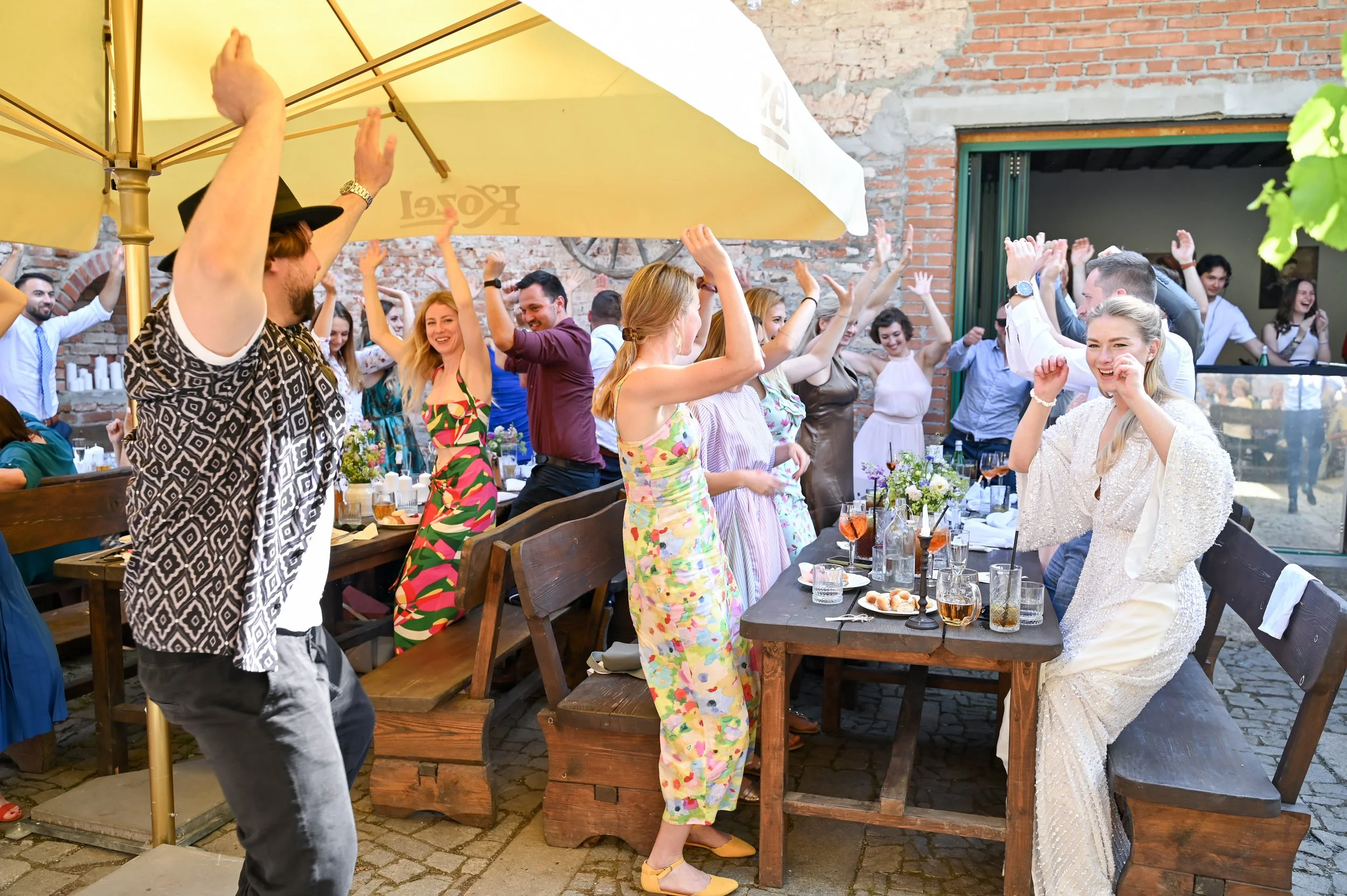 People celebrating and dancing outdoors at a party under a large yellow umbrella with food and drinks on the table.