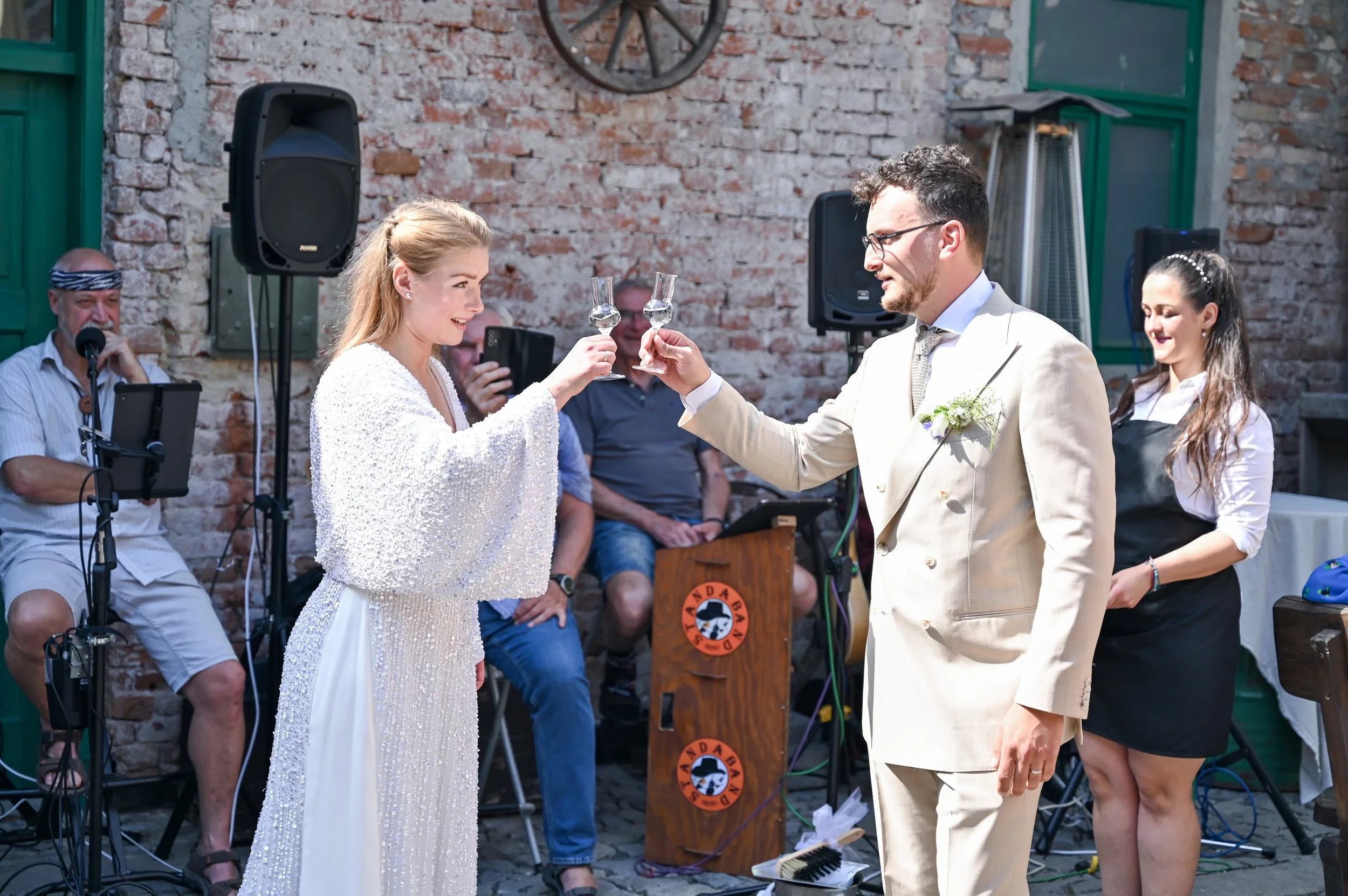 Bride and groom toasting with glasses during an outdoor wedding celebration. People in the background, microphone, speakers, and a brick wall with a wagon wheel are visible.