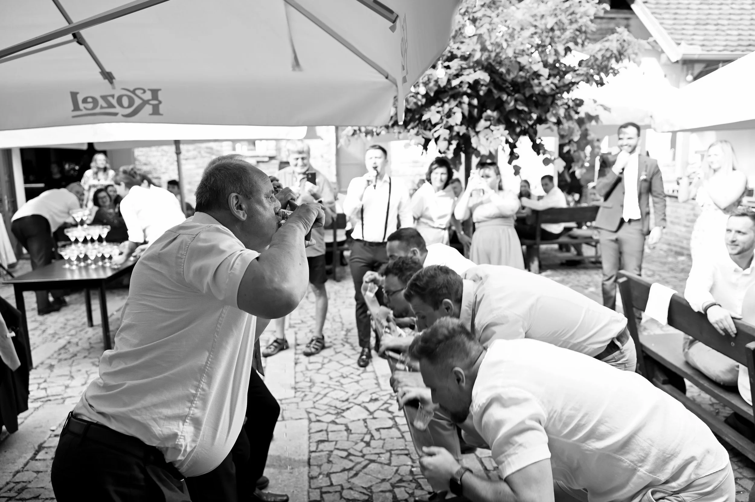 A black and white photo of a group of people at an outdoor gathering, where some are bending forward to drink from glasses, while others stand and watch, under large umbrellas and trees.