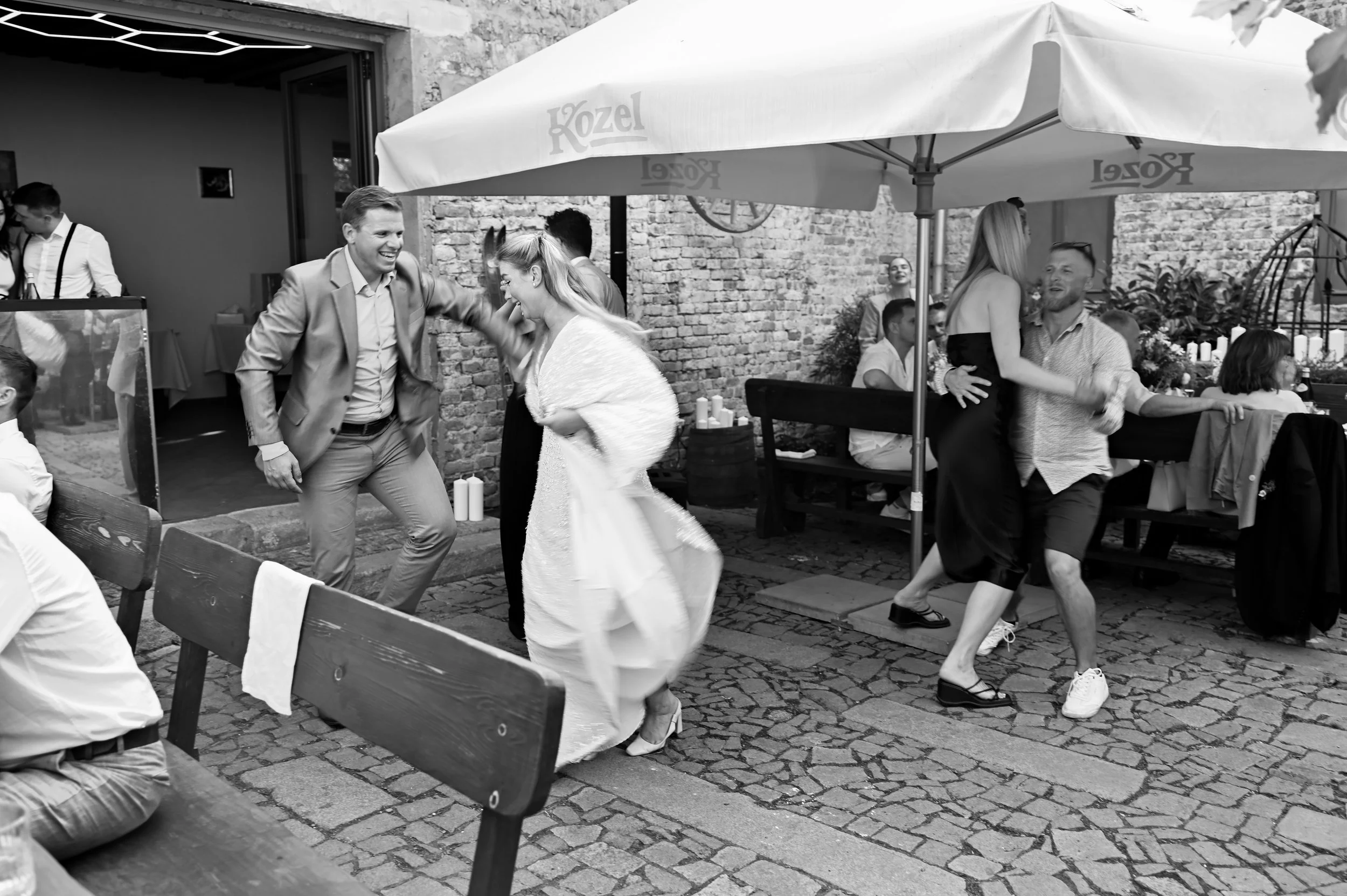 People dancing at an outdoor party or celebration under a large umbrella, with some seated at tables and others dancing on cobblestone ground.