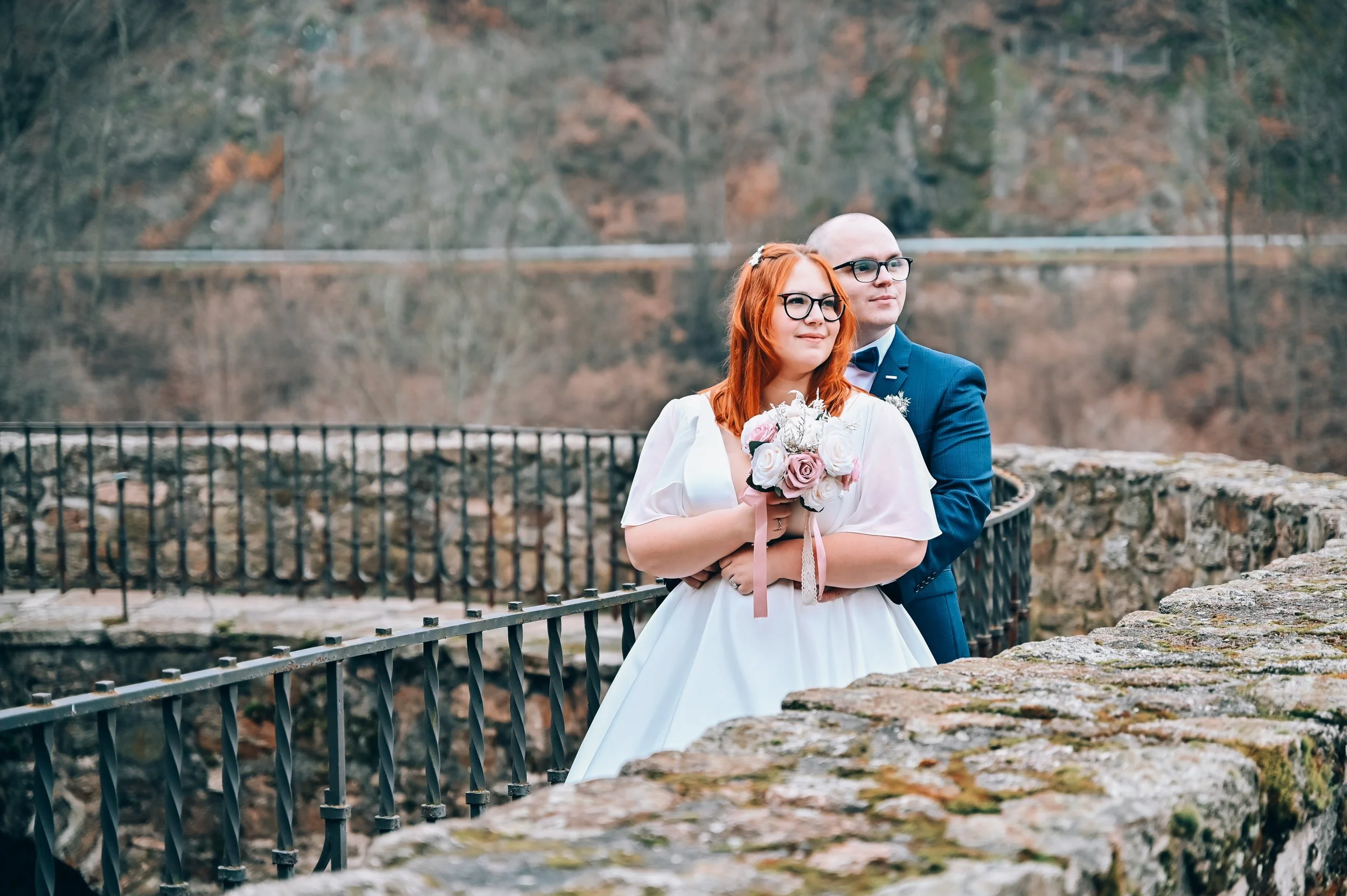 A bride and groom on a bridge outdoors, the bride holding a bouquet of pink and white roses, and the groom standing behind her, both looking off into the distance.