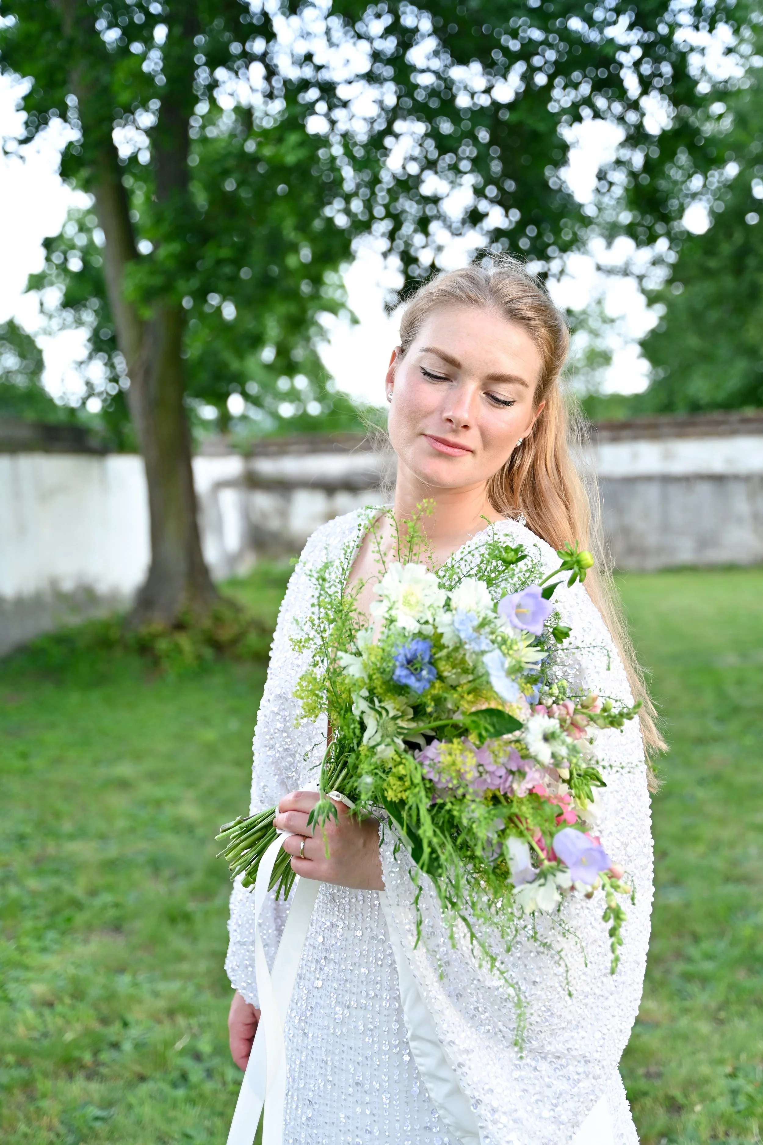 A woman in a white dress holding a colorful bouquet of flowers outdoors with trees and a fence in the background.