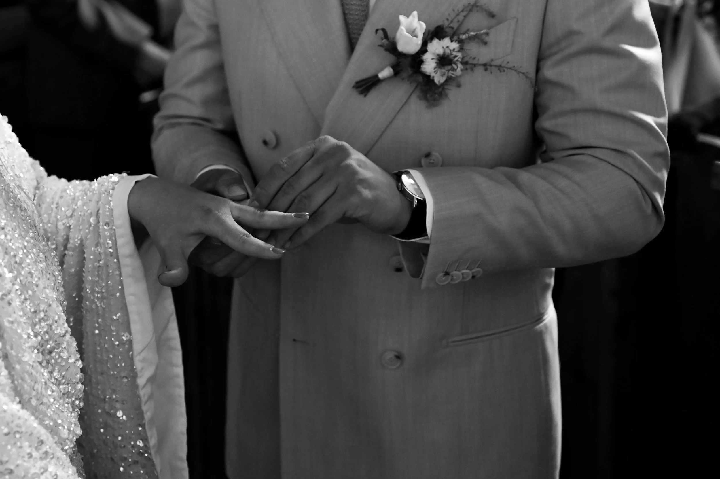 A groom holding a bride's hand during a wedding ceremony, both dressed in formal attire, with a focus on their hands and the groom's suit and boutonniere.
