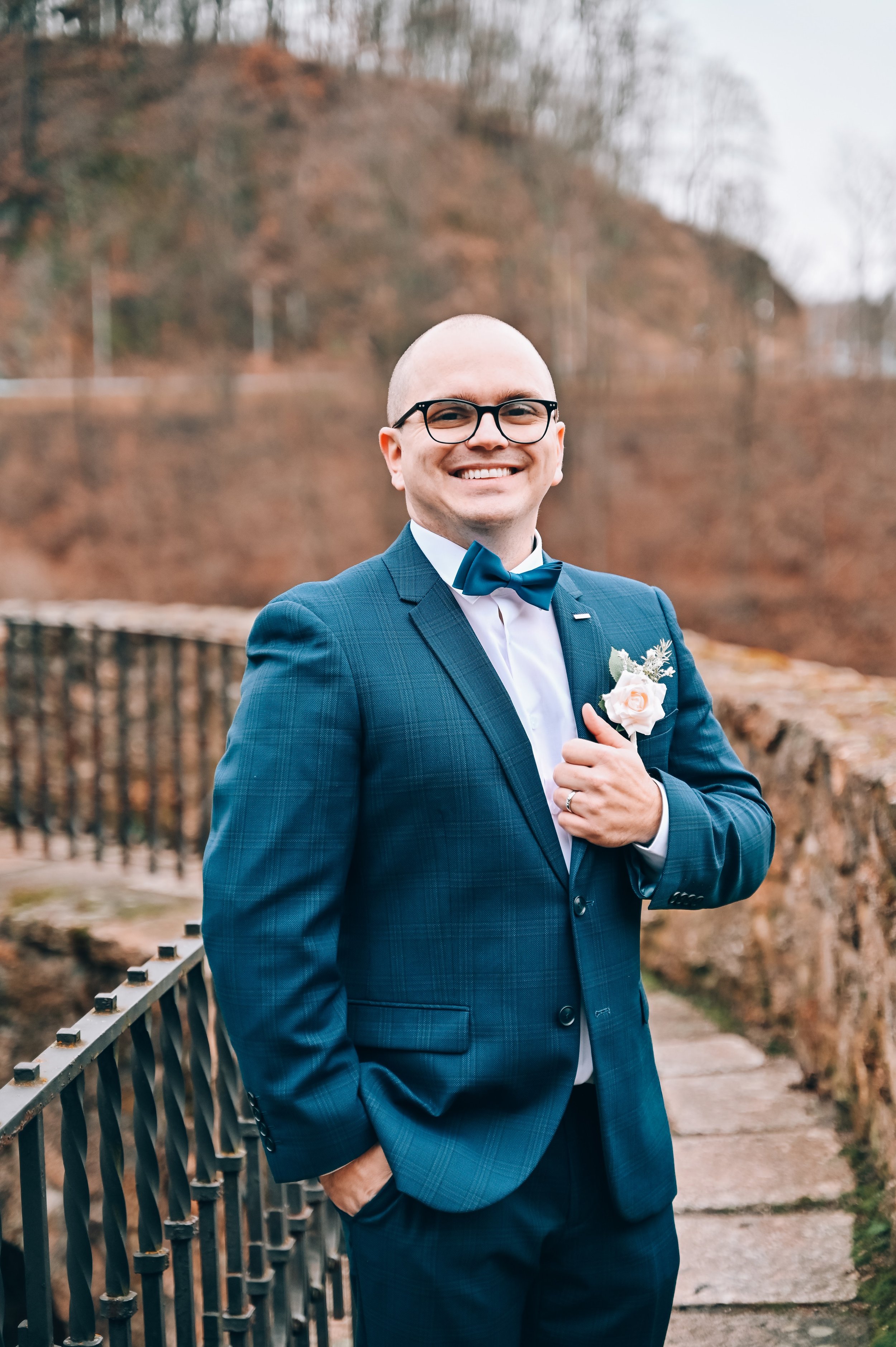 A man in a blue suit with a bow tie, wearing glasses, stands outdoors with one hand in his pocket and the other adjusting his shirt, smiling, with autumn foliage in the background.