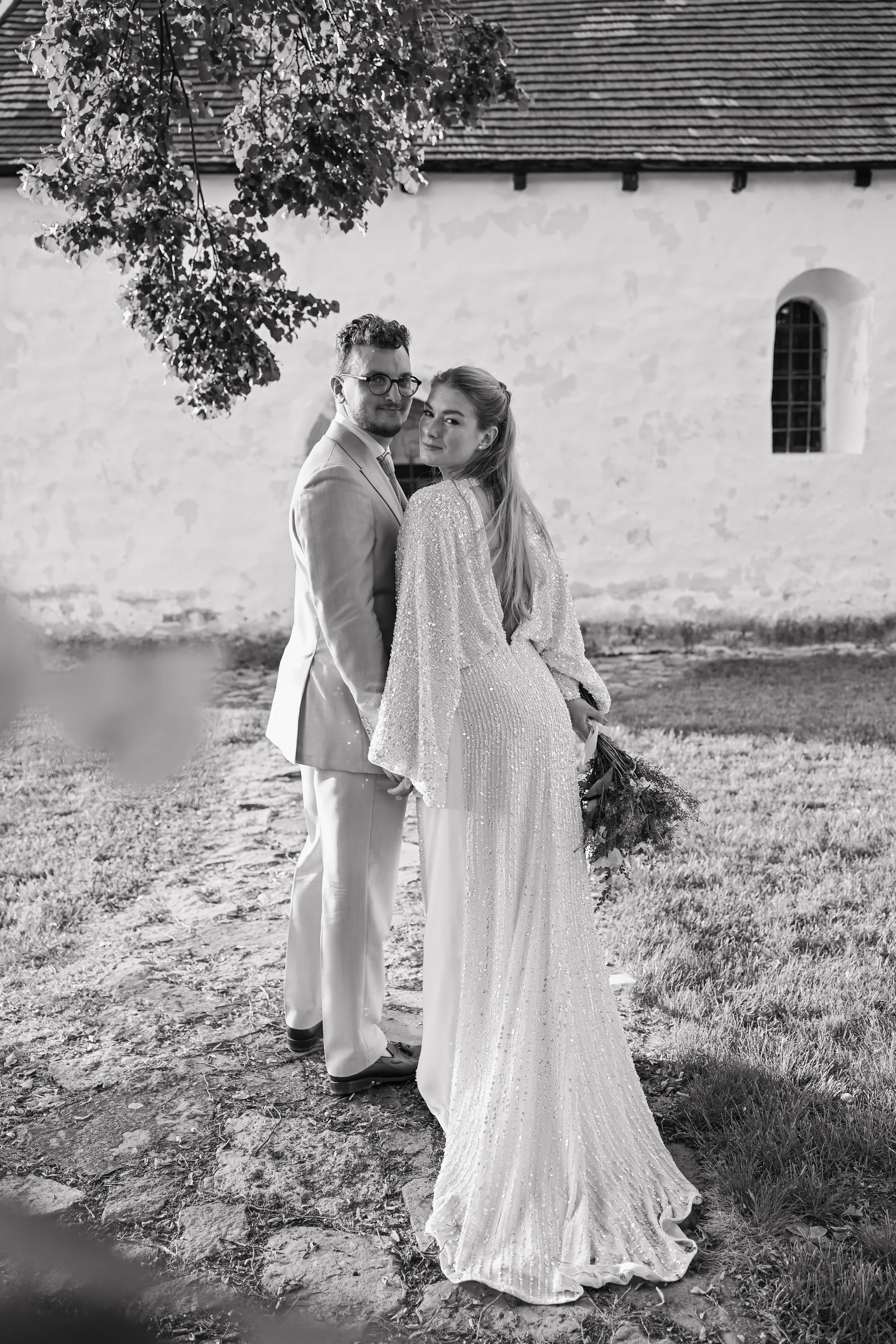 A black and white photo of a bride and groom standing outdoors on a dirt path in front of an old building, holding hands and looking at the camera, with a large tree branch overhead.