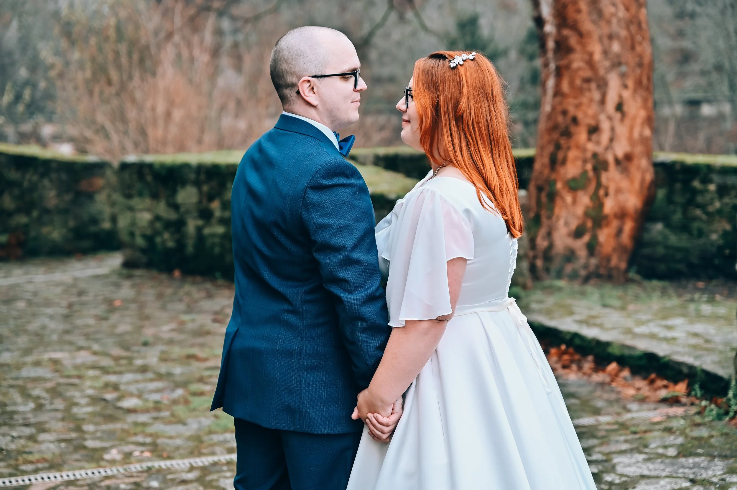 A couple dressed in wedding attire holding hands and gazing into each other's eyes outdoors around autumn, with trees and moss-covered stone structures in the background.