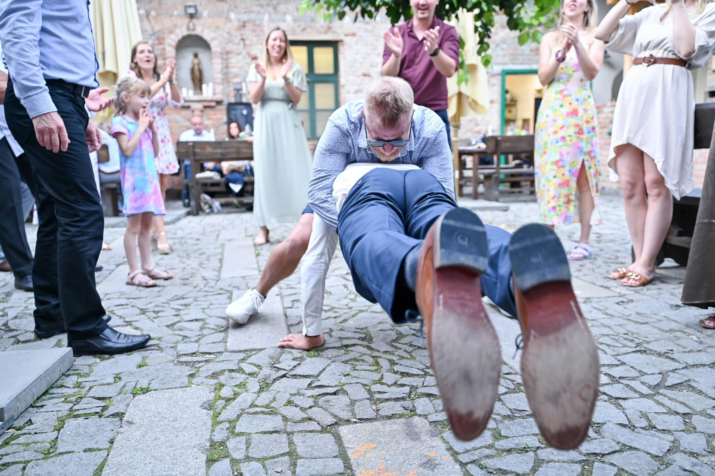 A man is being playfully flipped upside down on a cobblestone patio during a celebration, surrounded by smiling and clapping guests of various ages, with a brick building and greenery in the background.