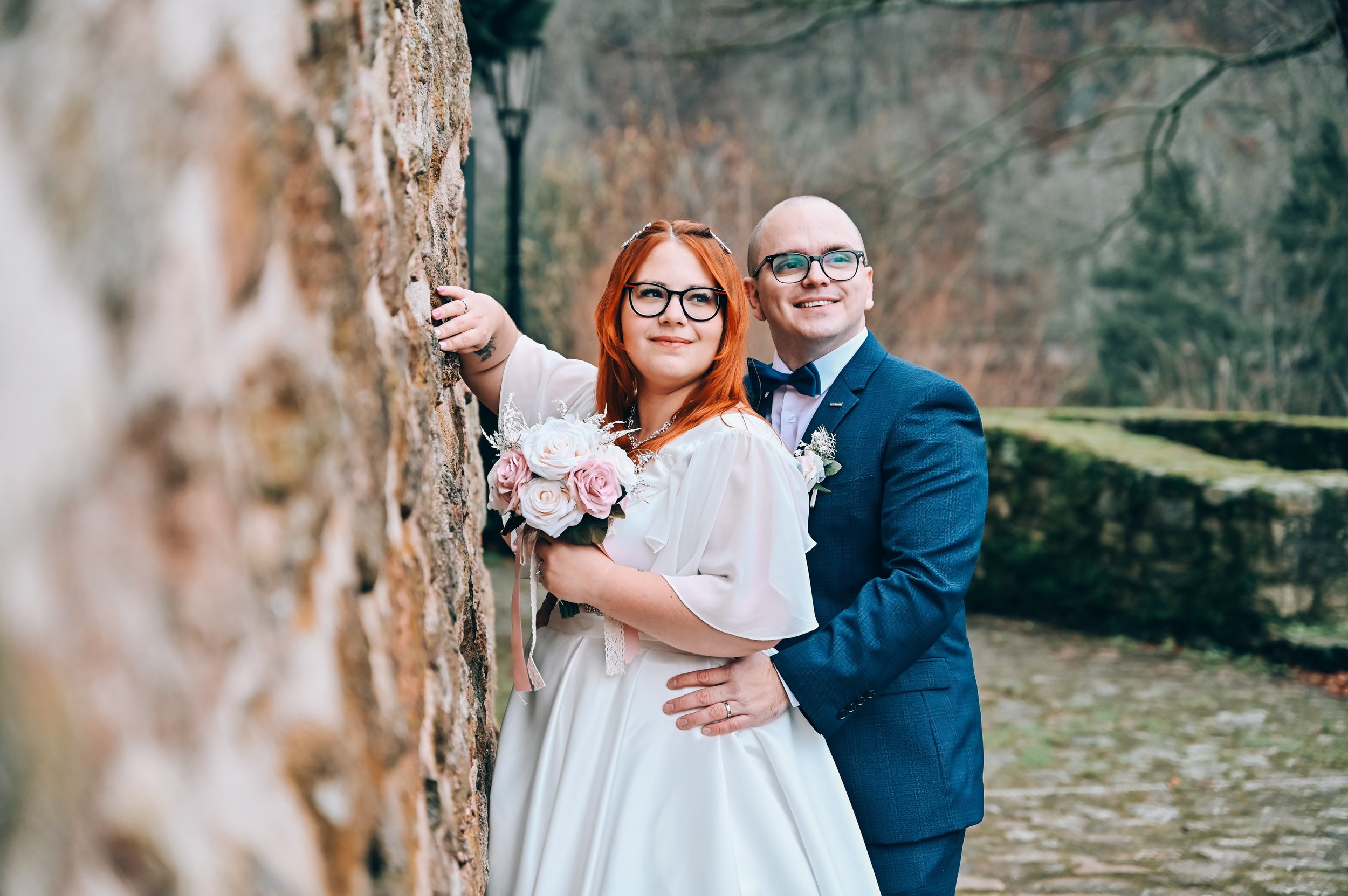 A bride and groom in wedding attire standing outdoors near a stone wall, with the bride holding a bouquet of pink and white roses, both smiling and looking at the camera.