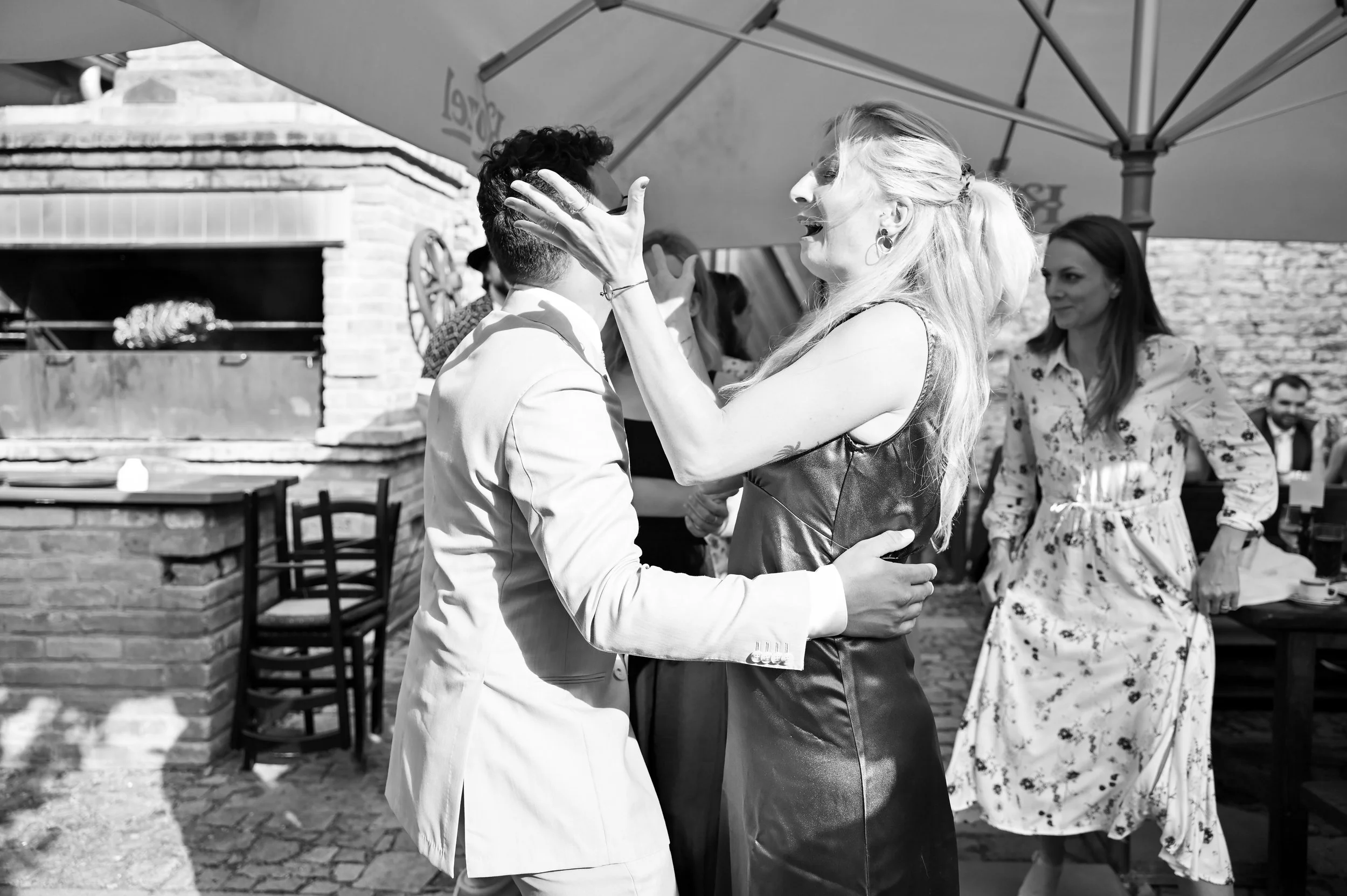 A woman is touching the face of a man, and they appear to be dancing or embracing outdoors under an umbrella, with other people in the background.