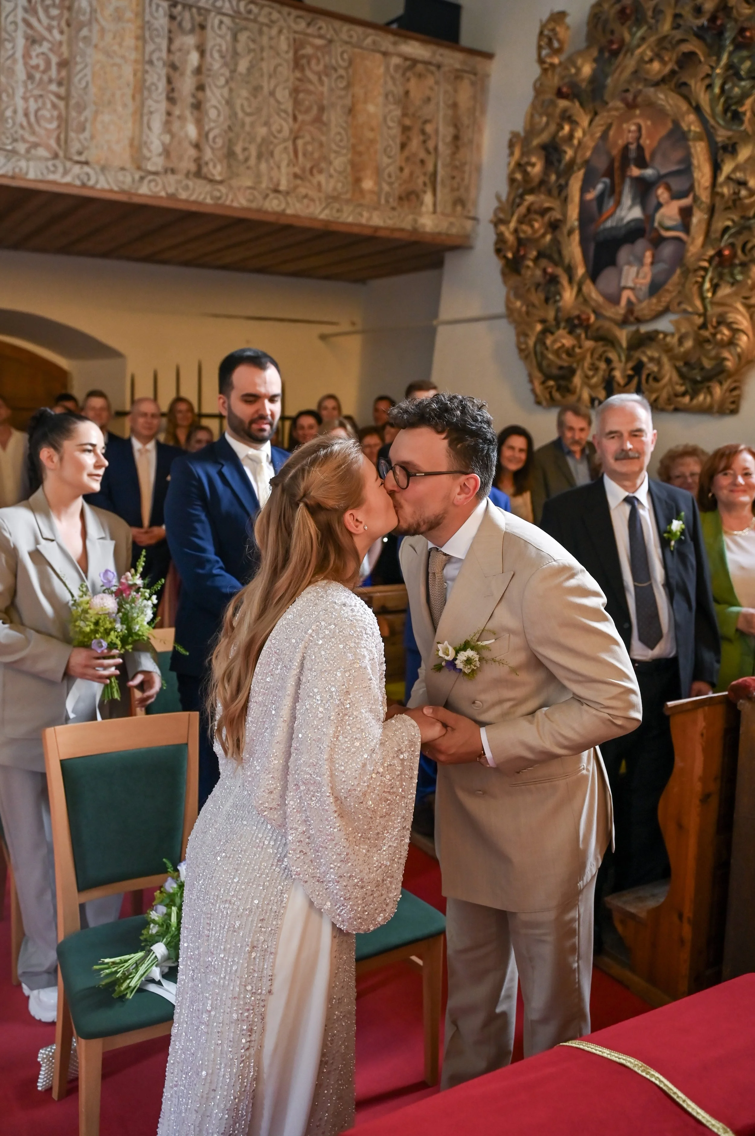 A bride and groom kissing during their wedding ceremony inside a church, surrounded by friends and family.