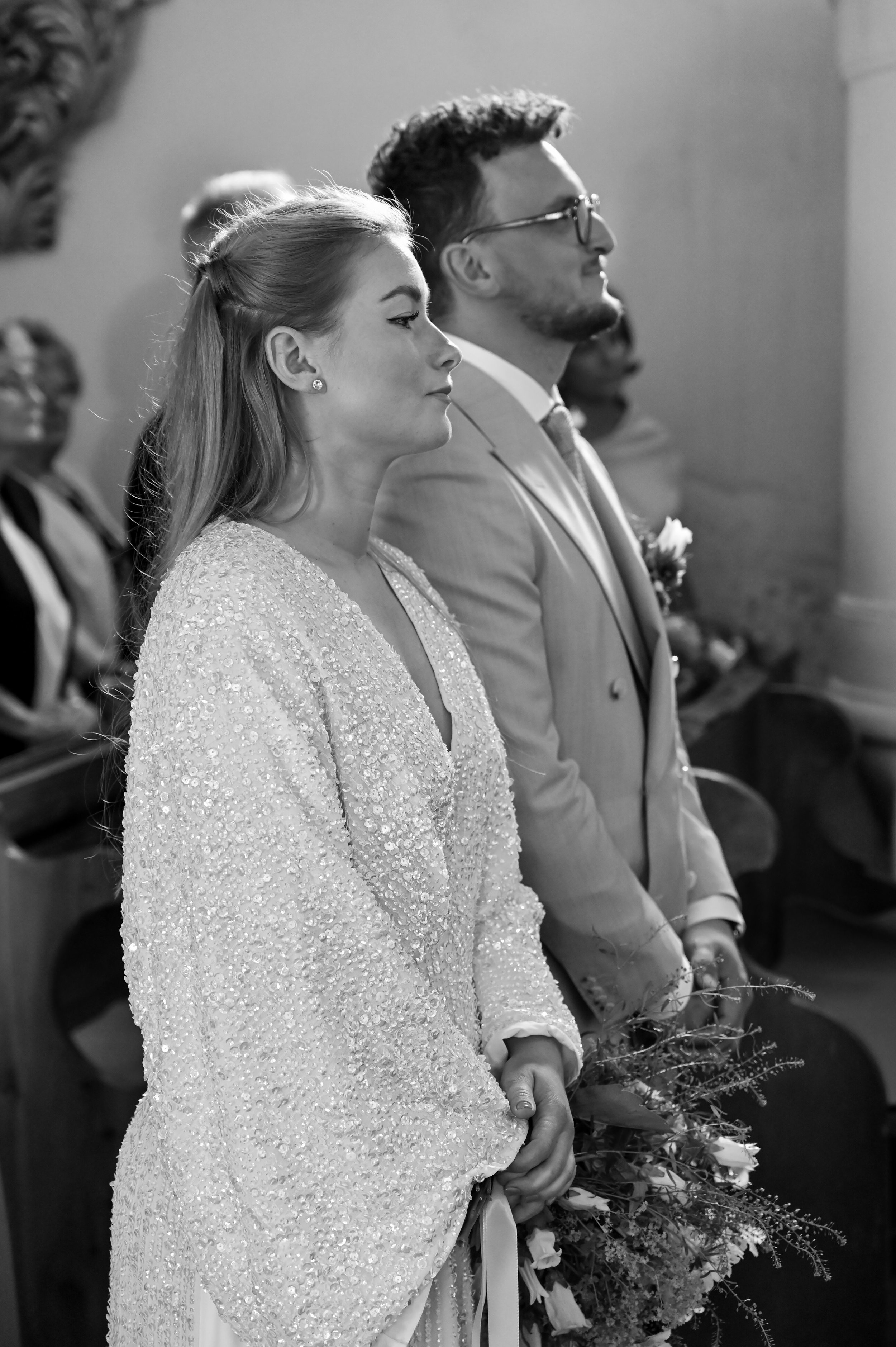 Black and white photo of a wedding ceremony showing a bride and groom standing side by side in profile, holding flowers, with guests in the background.