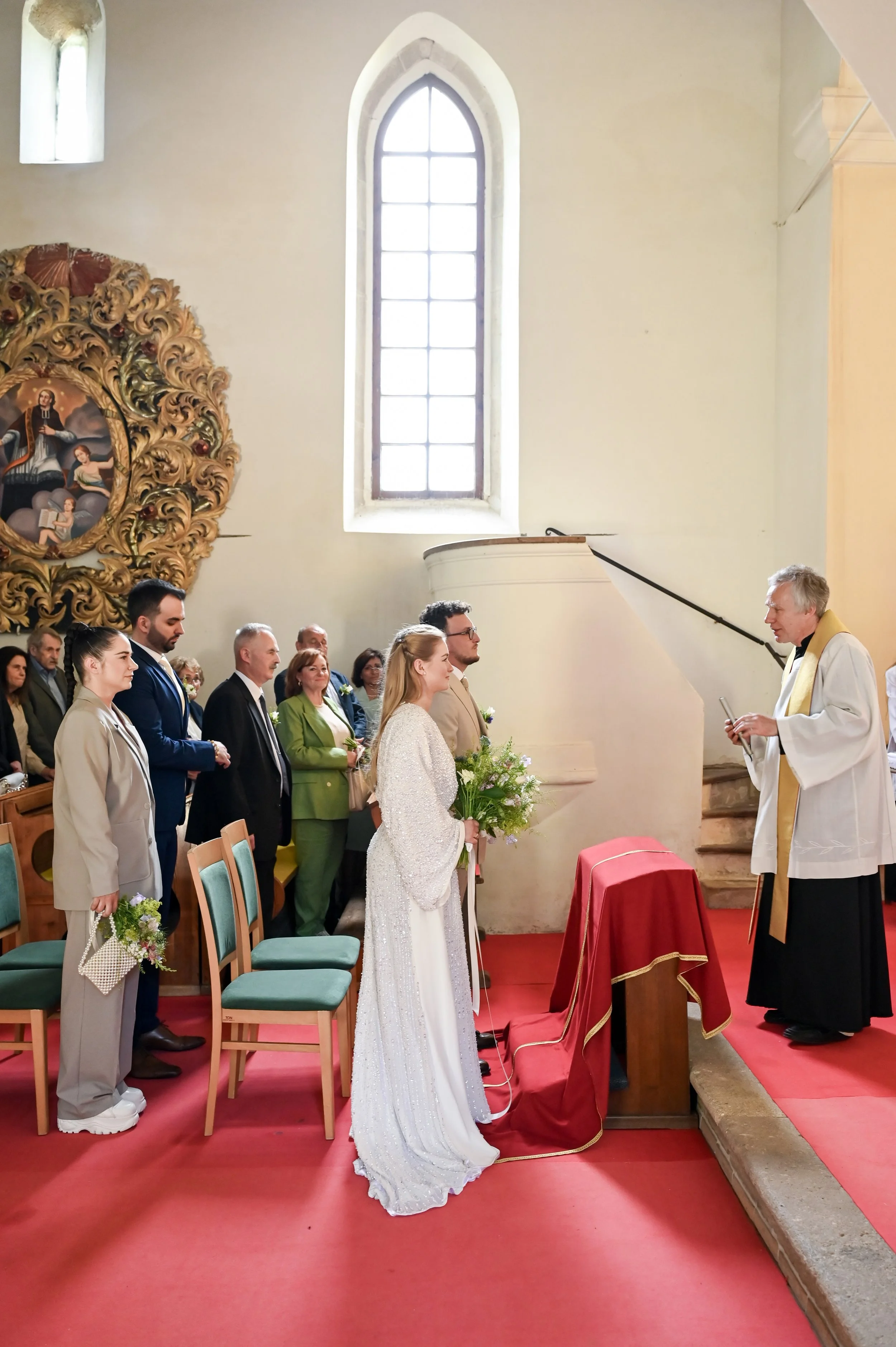 A wedding ceremony taking place inside a church, with a priest officiating. The bride and groom are standing in front of the priest, holding flowers, with guests seated behind them. The church has large windows and ornate decorations on the wall.